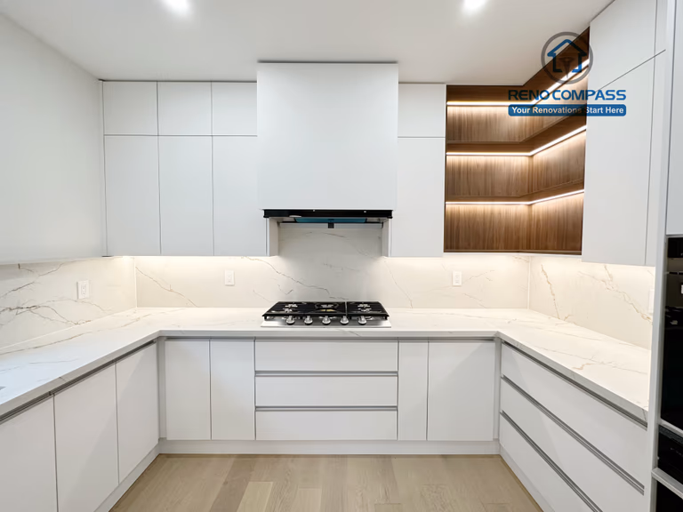 This image showcases a modern U-shaped kitchen renovation in Richmond Hill, featuring full-height custom white cabinetry, a centrally positioned gas cooktop, and seamless quartz countertops with a marble-look backsplash. The clean, handle-less cabinet design maximizes storage and workflow efficiency, while recessed ceiling lighting provides even illumination throughout the space. Warm wood accent shelving with integrated LED lighting adds contrast and depth. Completed by a professional Toronto Renovation Company, this kitchen renovation emphasizes smart layout planning, functionality, and contemporary craftsmanship.
