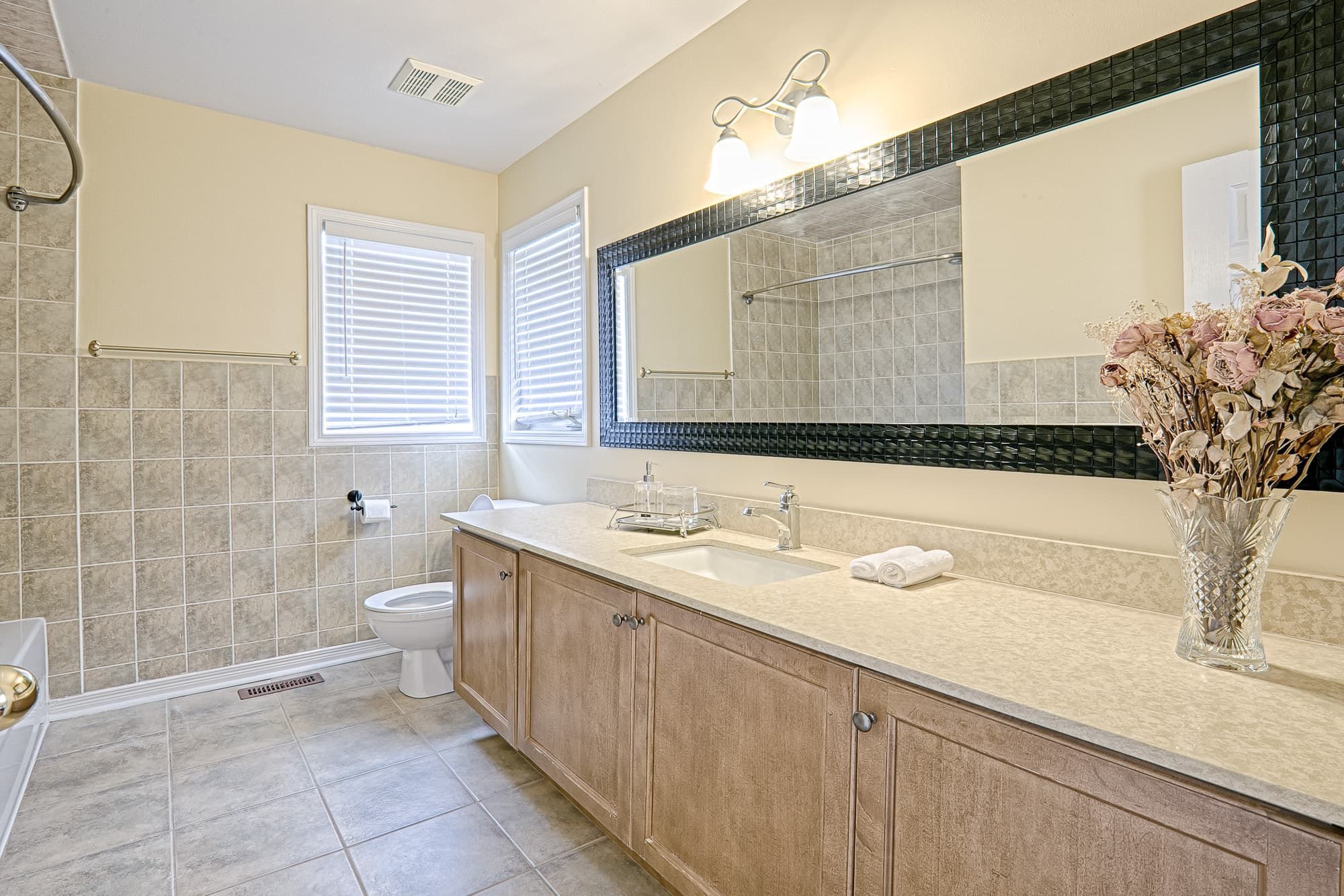 This bright bathroom features a simple and lovely interior design with a spacious double vanity, neutral tile finishes, and a large framed mirror that enhances depth and light. The warm wood cabinetry paired with soft beige tones creates a clean and comfortable atmosphere. This bathroom design focuses on functionality, storage efficiency, and everyday comfort, making it ideal for family homes and long-term residential use.