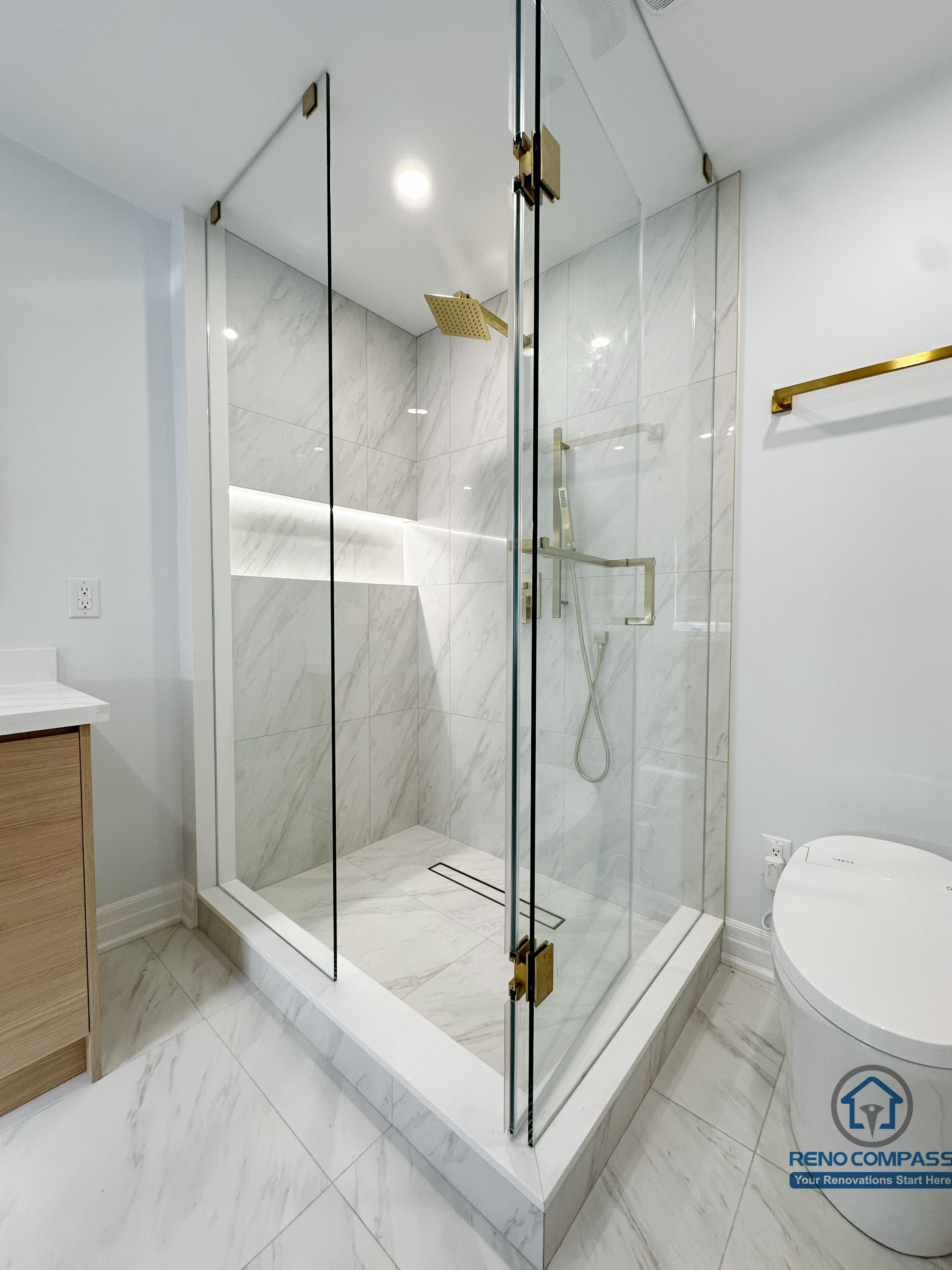 Modern walk-in shower featuring full-height glass enclosure, marble-look wall tiles, integrated shower niche lighting, and brushed gold fixtures, creating a clean, elegant, and highly functional bathroom layout.