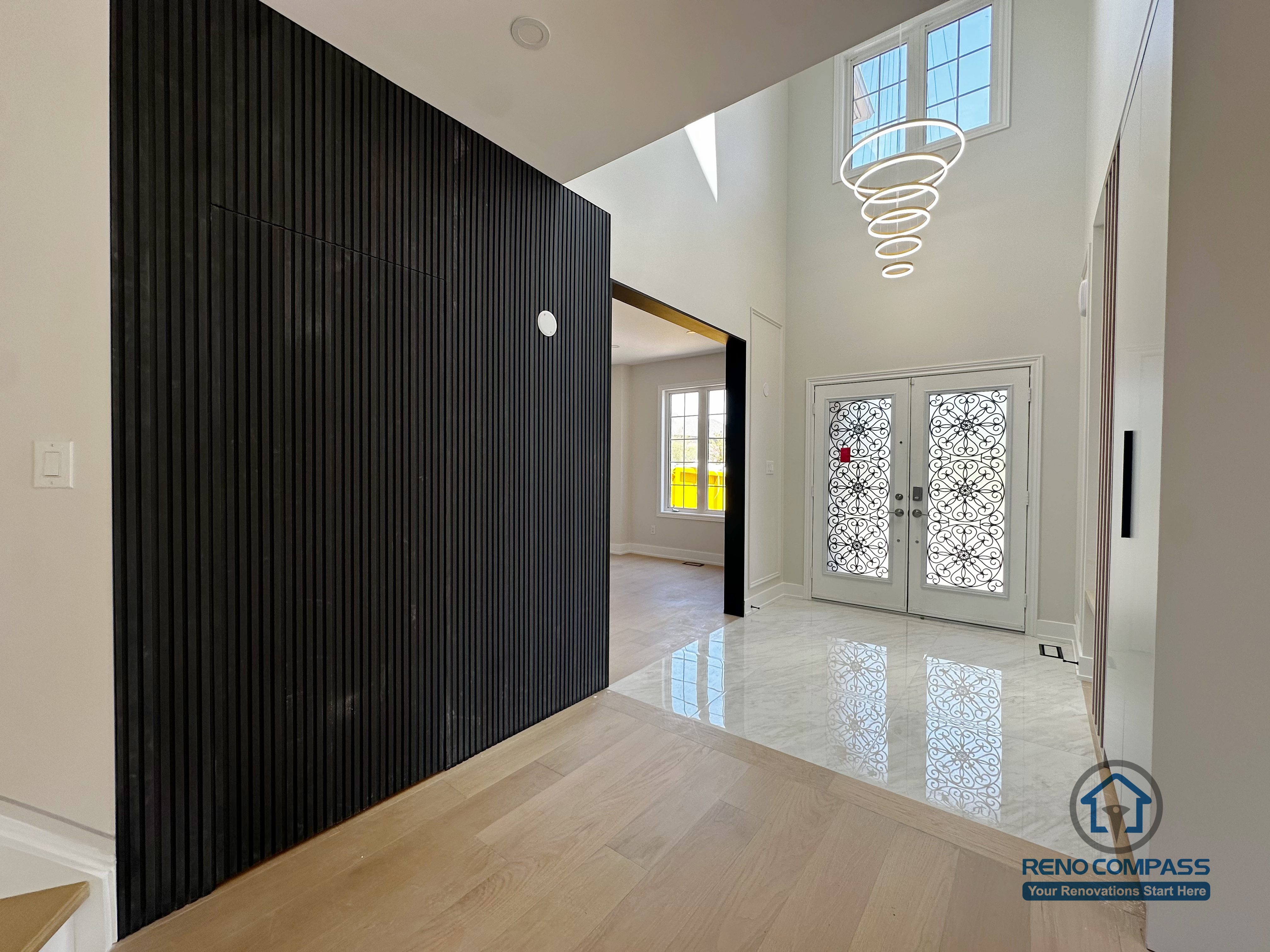 This image highlights the entryway from a Markham interior renovation, where architectural contrast and material layering were used to create a strong first impression. A full-height vertical slat feature wall in a dark finish defines the foyer zone, adding depth and visual rhythm while subtly separating the entry from the main living areas.

The double-height ceiling allows natural light to flow through the space, complemented by a modern sculptural chandelier that emphasizes vertical openness. Polished flooring near the entrance transitions seamlessly into light wood flooring, balancing elegance with everyday practicality.

Decorative double front doors introduce both privacy and natural light, reinforcing the refined yet welcoming character of the home. This entryway renovation reflects Reno Compass’s approach to interior design—using proportion, lighting, and material contrast to elevate first impressions in modern Markham homes.