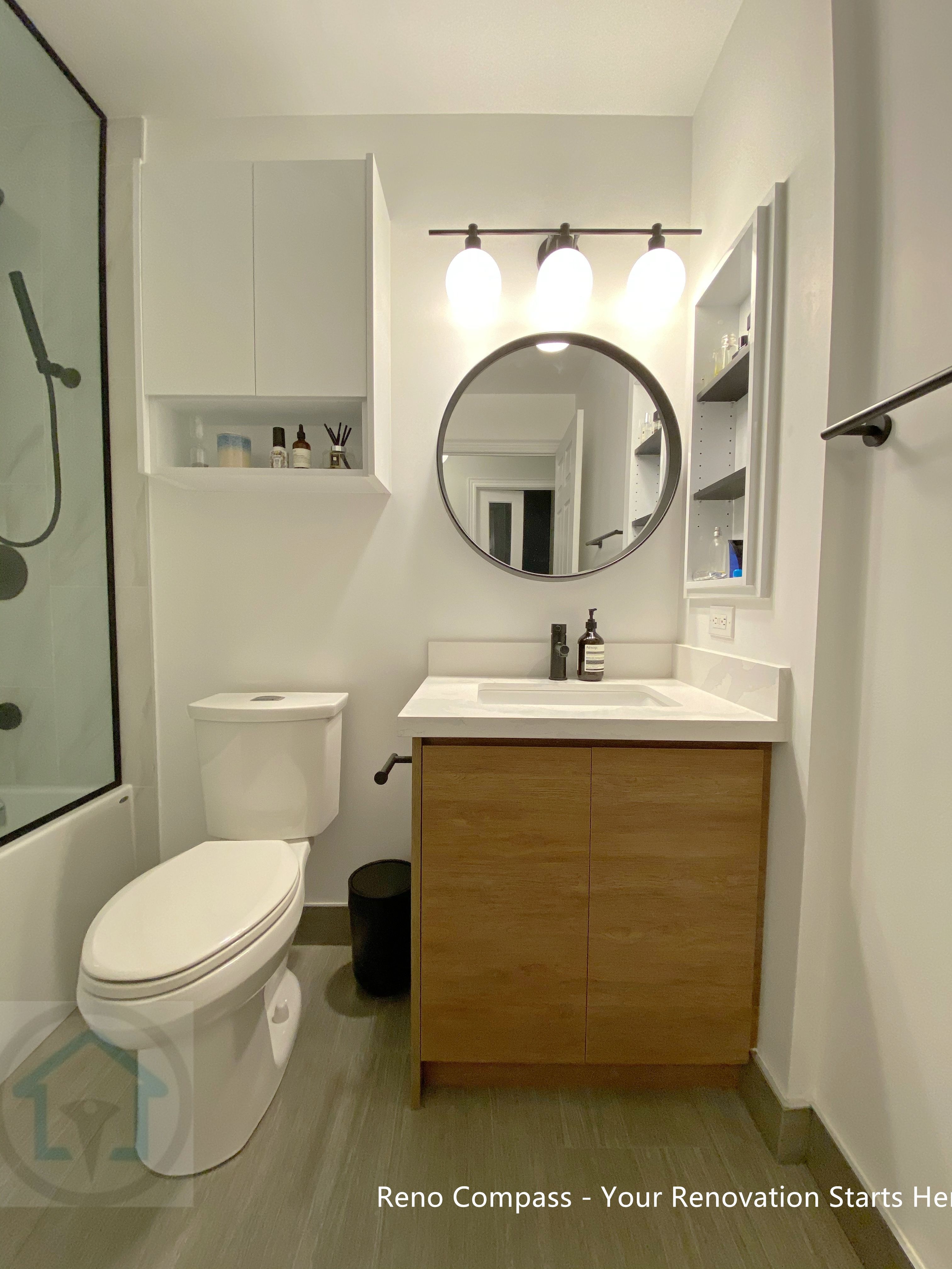 This image features a modern bathroom renovation in a North York condo, highlighting a floating wood-tone vanity paired with a round mirror and wall-mounted lighting. The layout combines a compact bathtub-shower enclosure with clean-lined fixtures and built-in storage niches, maximizing functionality within a limited footprint. Neutral finishes and balanced proportions create a calm, contemporary bathroom designed for everyday comfort and long-term use.