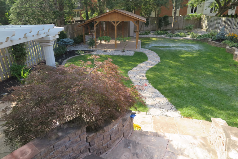 This image presents a full view of a North York landscaping renovation featuring a curved natural stone walkway, manicured lawn, raised garden beds, and a wooden pavilion as the focal point. The design creates smooth circulation, clear outdoor zoning, and a balanced connection between planting, hardscape, and lawn areas. Completed by a Toronto Renovation Company specializing in custom residential landscaping and backyard transformations.