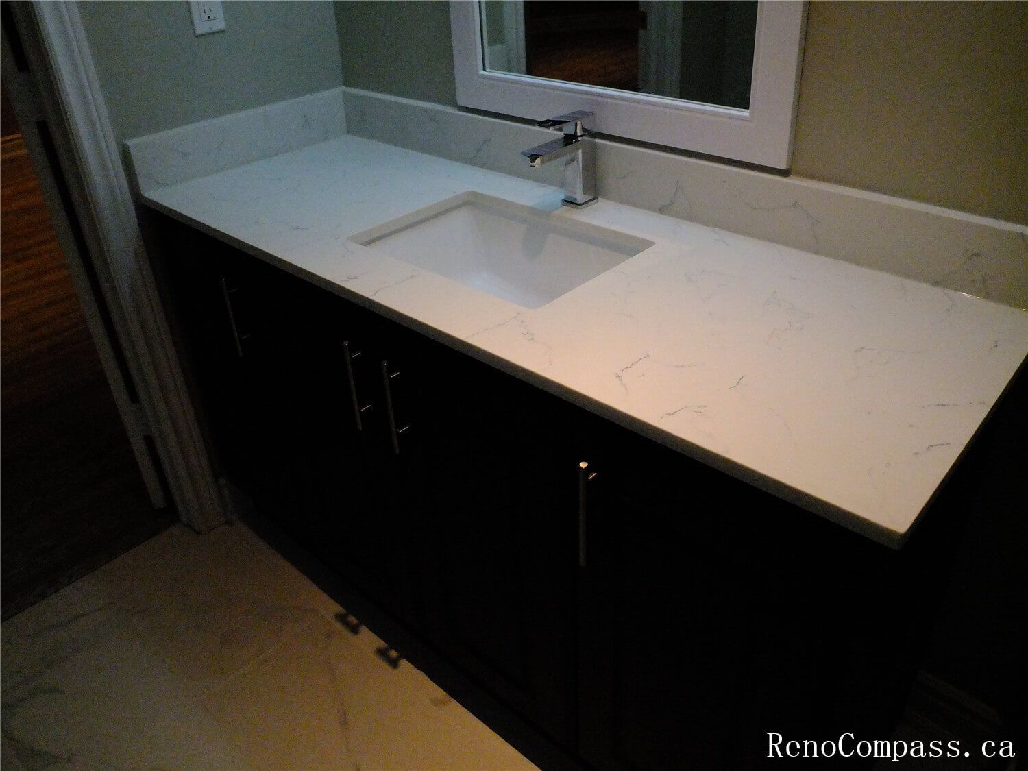Close-up view of the bathroom vanity installed in this Ajax basement renovation, featuring a sleek quartz countertop with an integrated sink, modern black faucet, and dark storage cabinetry. The large mirror enhances brightness while the curved glass shower enclosure complements the contemporary bathroom design. Completed by a professional Toronto Renovation Company, this basement bathroom upgrade combines elegant finishes with practical functionality.