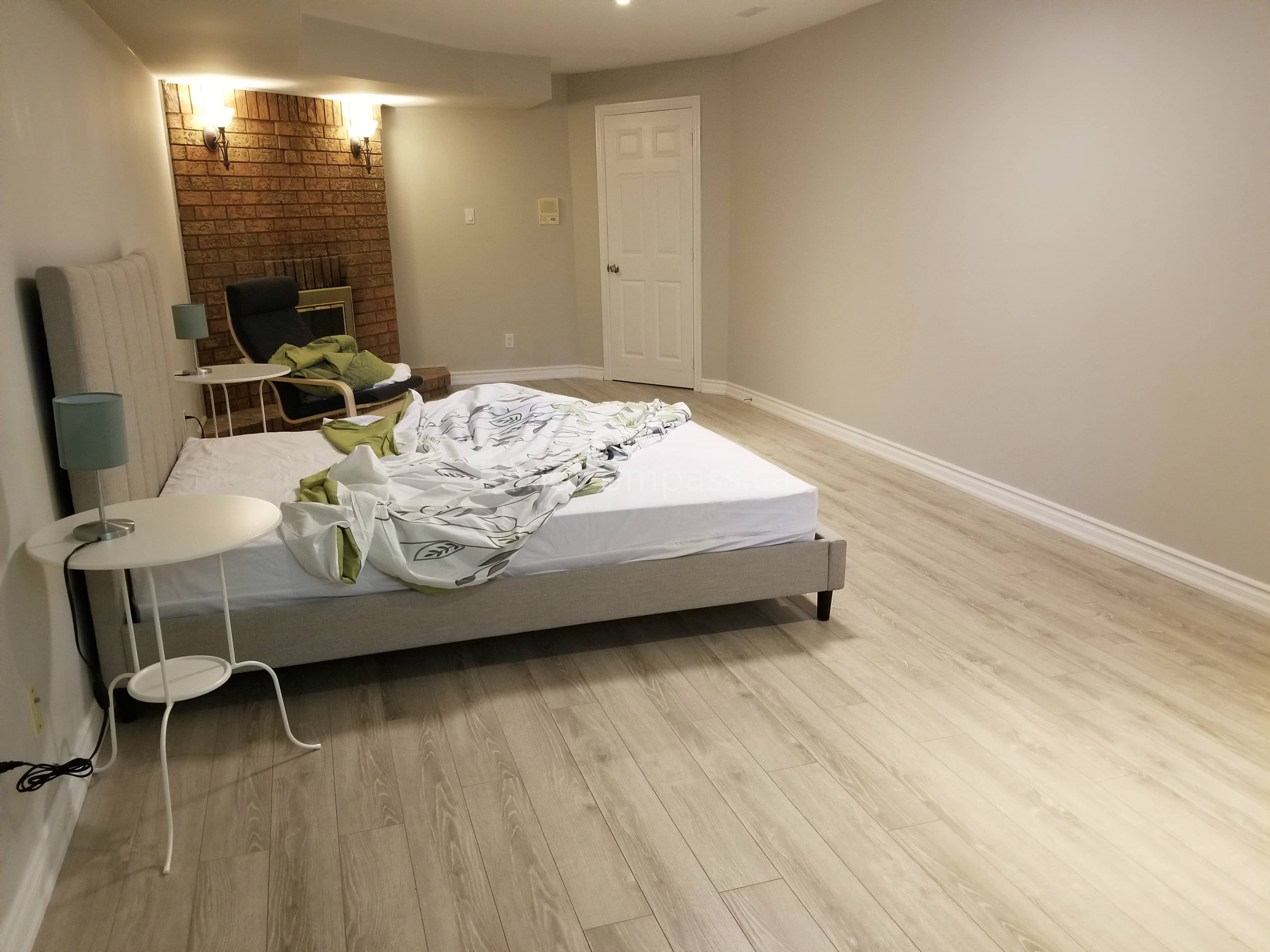 This Toronto basement renovation features a bright and spacious bedroom area with newly installed light wood flooring, fresh neutral wall paint, and modern baseboards. The open layout includes a comfortable sleeping area and workspace, creating a functional and cozy basement living environment. Completed by Reno Compass Inc., this basement upgrade enhances comfort, style, and usability, making the space ideal for rental or family living.