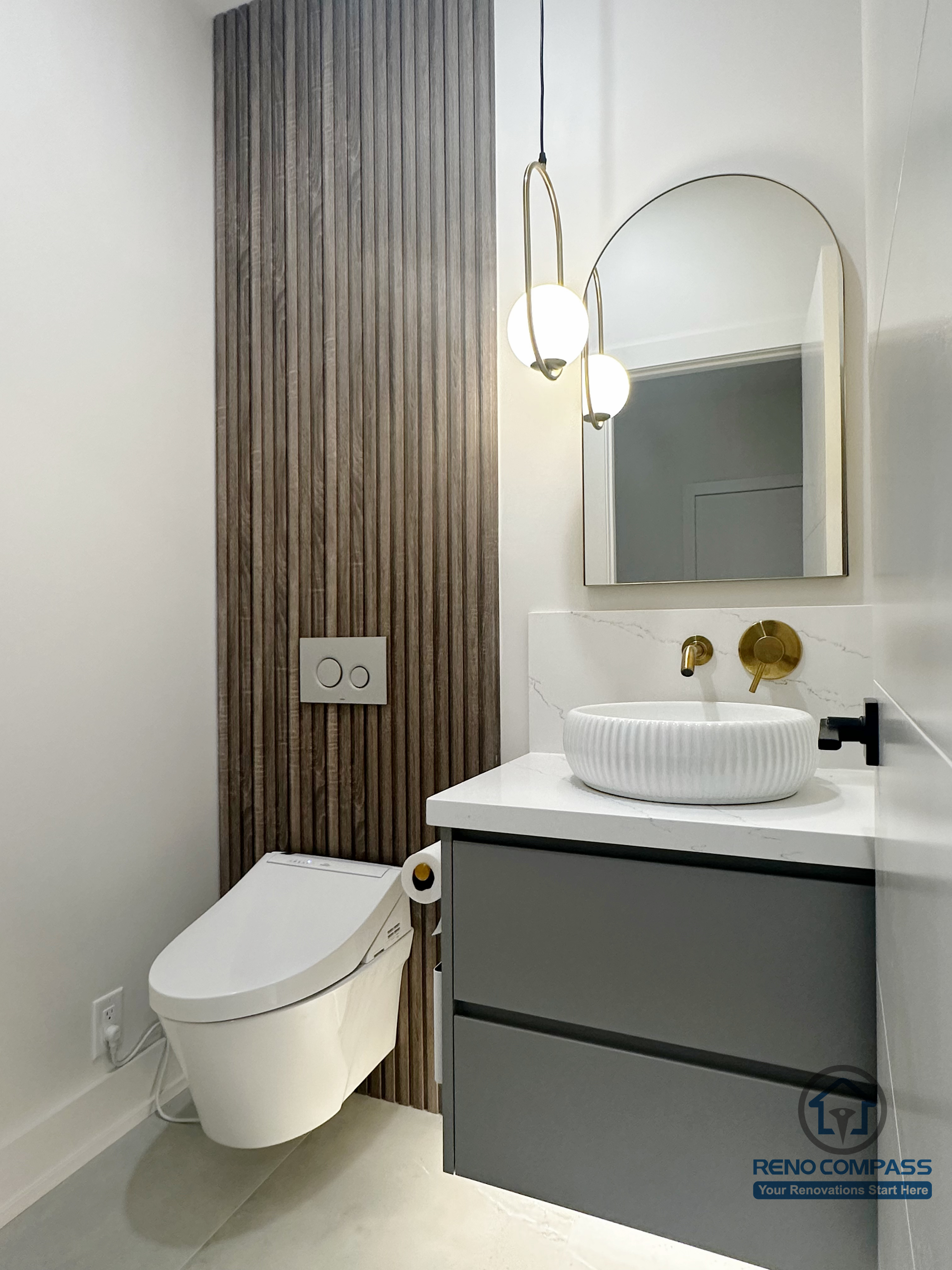 This Oakville powder room renovation highlights a floating grey vanity paired with a vessel sink and warm wood accent wall. Brushed brass fixtures, soft pendant lighting, and clean lines create a refined, modern space that balances visual warmth with everyday functionality.