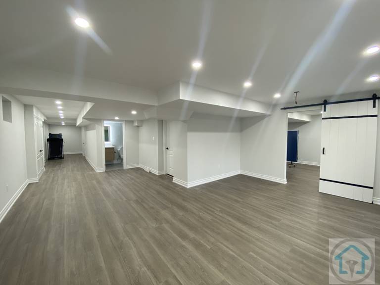 This Aurora basement renovation features an open-concept layout with modern flooring, bright recessed lighting, and clean white walls. The design maximizes natural light and creates a versatile living space suitable for family activities or entertainment. Contractor: Reno Compass Inc., City: Aurora.