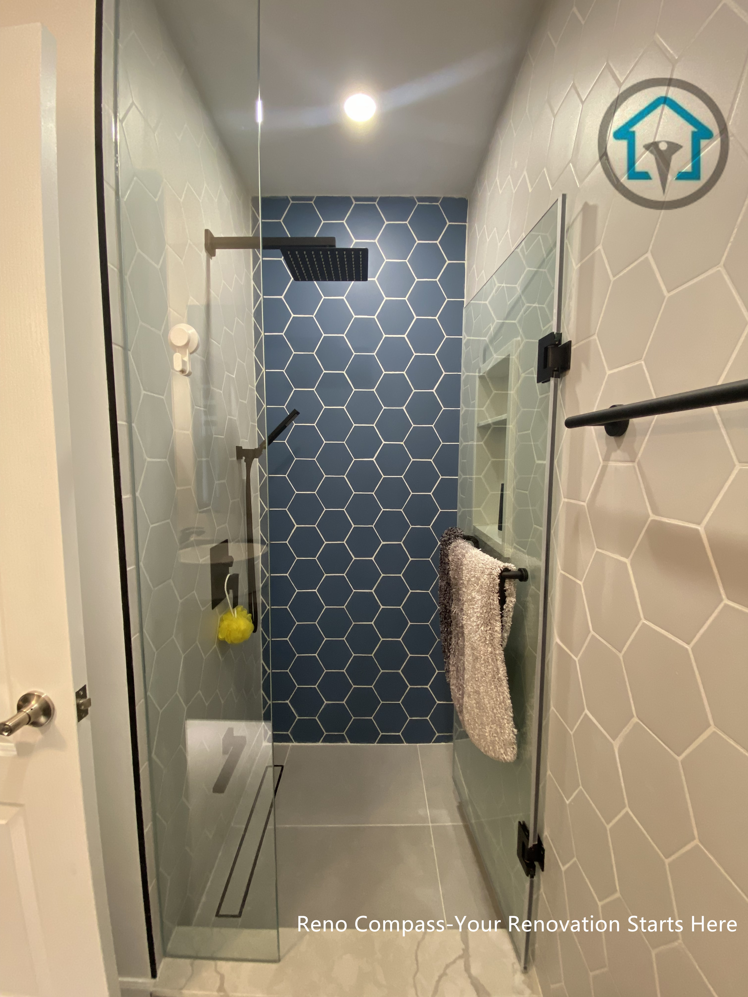 This bathroom renovation in Toronto features a walk-in shower highlighted by a blue hex tile accent wall that adds depth and visual interest to the space. Frameless glass panels keep the layout feeling open, while matte black fixtures provide a clean contrast against the light-toned wall tiles. A linear drain and compact proportions make the shower both functional and easy to maintain, demonstrating a thoughtful balance between design detail and everyday practicality.