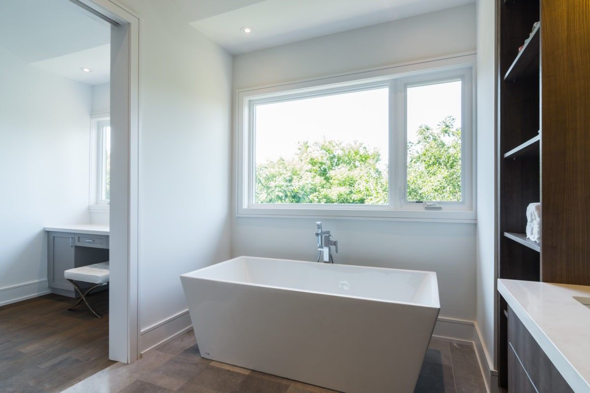 This Toronto modern bathroom renovation features a sleek freestanding bathtub positioned beside a large window, allowing natural light to enhance the clean and relaxing atmosphere. The space is complemented by custom cabinetry, a minimalist vanity area, and warm wood flooring, creating a spa-like environment with both functionality and elegance.