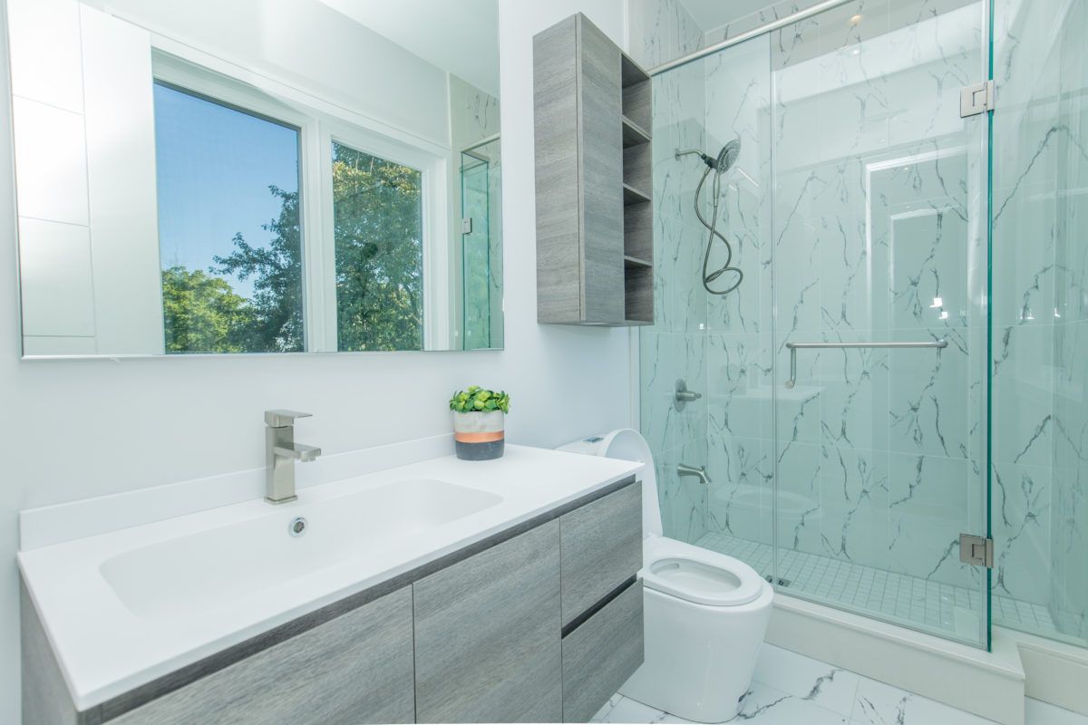 Bright modern bathroom renovation in a 5000 sqft Toronto home featuring a frameless glass shower enclosure, marble-style wall tiles, floating grey vanity with integrated sink, and large mirror reflecting natural light. The clean layout and contemporary finishes create a spacious spa-like atmosphere with functional storage and elegant detailing. Completed by Reno Compass Inc., a professional Toronto renovation company specializing in high-end bathroom and full home interior renovations.