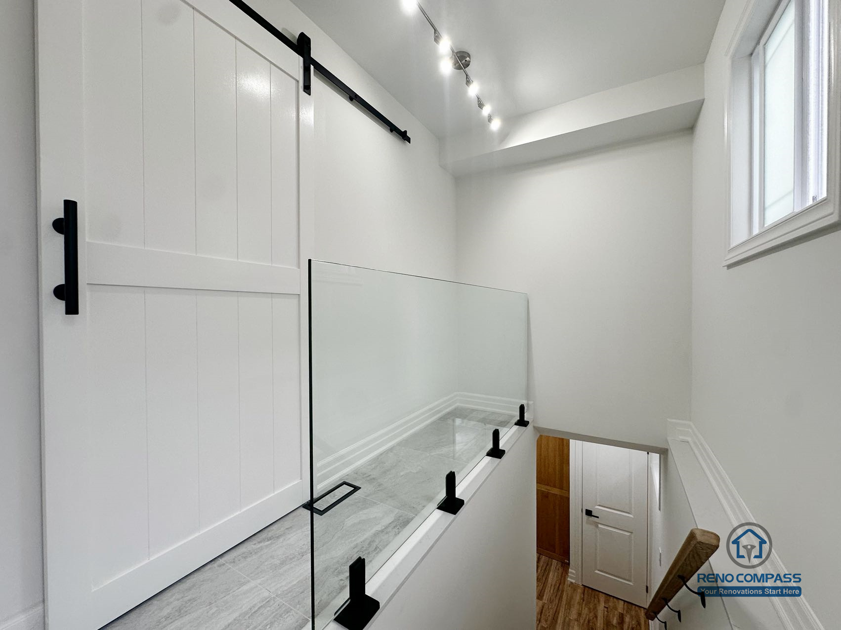 This image presents a horizontal view of the basement stair landing in a completed Pickering basement renovation, emphasizing safety, visibility, and natural light. A glass guardrail provides secure edge protection while maintaining visual openness across the stairwell. The landing is finished with durable tile flooring, creating a clear and practical transition zone between levels. Track lighting and a nearby window enhance brightness and usability, supporting safe movement throughout the space. Clean finishes and restrained detailing allow this transitional area to integrate seamlessly with the overall basement layout.