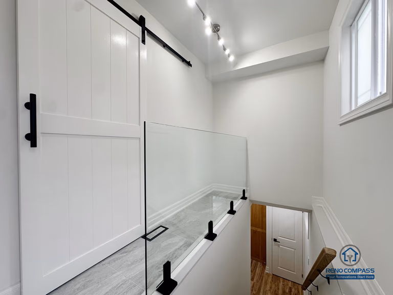 This image presents a horizontal view of the basement stair landing in a completed Pickering basement renovation, emphasizing safety, visibility, and natural light. A glass guardrail provides secure edge protection while maintaining visual openness across the stairwell. The landing is finished with durable tile flooring, creating a clear and practical transition zone between levels. Track lighting and a nearby window enhance brightness and usability, supporting safe movement throughout the space. Clean finishes and restrained detailing allow this transitional area to integrate seamlessly with the overall basement layout.
