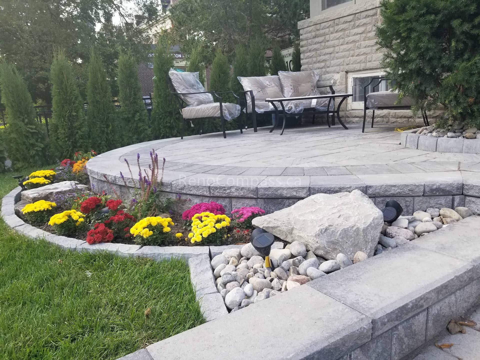 This Toronto landscaping renovation features a beautifully designed raised stone patio with a curved layout, creating a comfortable outdoor seating and relaxation space. The project includes decorative garden beds with vibrant seasonal flowers, natural stone accents, river rock drainage detail, and modern landscape lighting. Surrounded by privacy hedges and fresh lawn, this professionally completed Toronto backyard landscaping upgrade enhances both functionality and curb appeal while providing a stylish outdoor living environment.