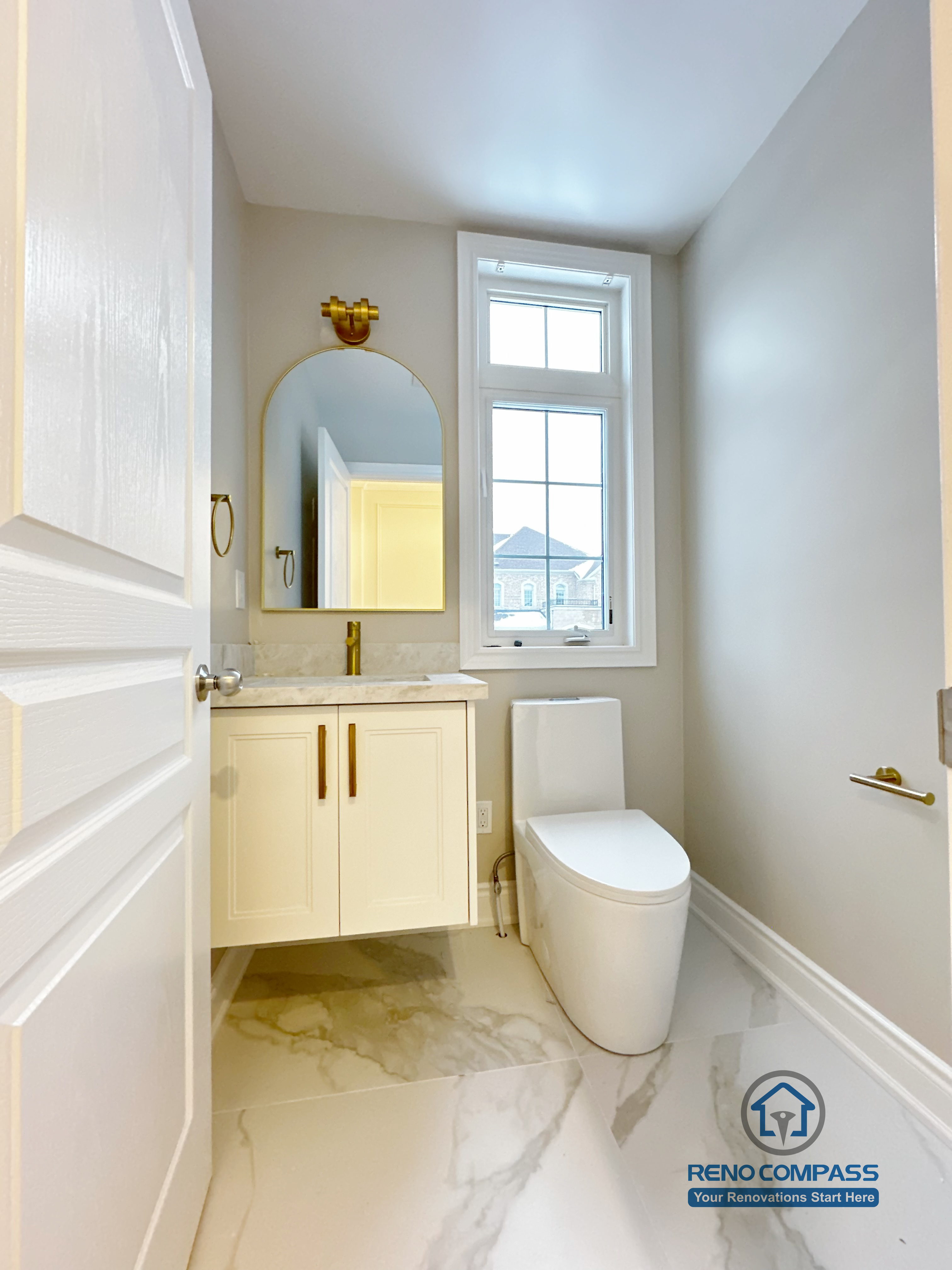 Private toilet area within a Vaughan primary bathroom renovation, featuring a compact custom vanity, arched mirror with gold accent lighting, modern toilet, and marble-look porcelain flooring. Designed to maximize natural light and privacy, this space was completed by a professional Toronto renovation company focused on efficient layout and cohesive design details.