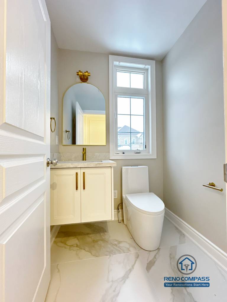 Private toilet area within a Vaughan primary bathroom renovation, featuring a compact custom vanity, arched mirror with gold accent lighting, modern toilet, and marble-look porcelain flooring. Designed to maximize natural light and privacy, this space was completed by a professional Toronto renovation company focused on efficient layout and cohesive design details.