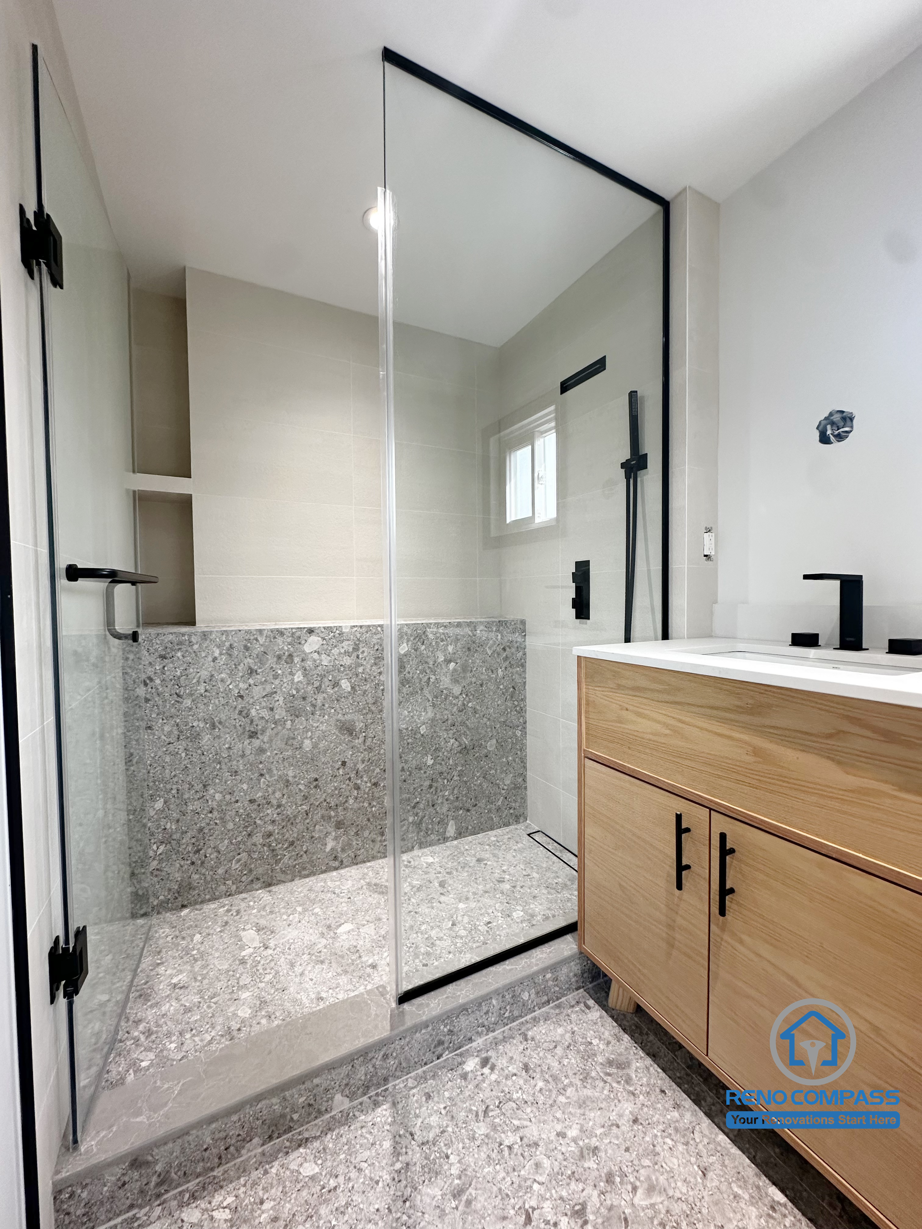 This view captures the connection between the walk-in shower and vanity area in a Richmond Hill bathroom renovation. The frameless glass shower, textured stone flooring, and warm wood-finish vanity create a balanced contrast between natural materials and clean modern lines. Matte black fixtures add definition, while the open layout enhances both light flow and usability. Designed and completed by Reno Compass, this project reflects a refined contemporary bathroom renovation in the Greater Toronto Area.