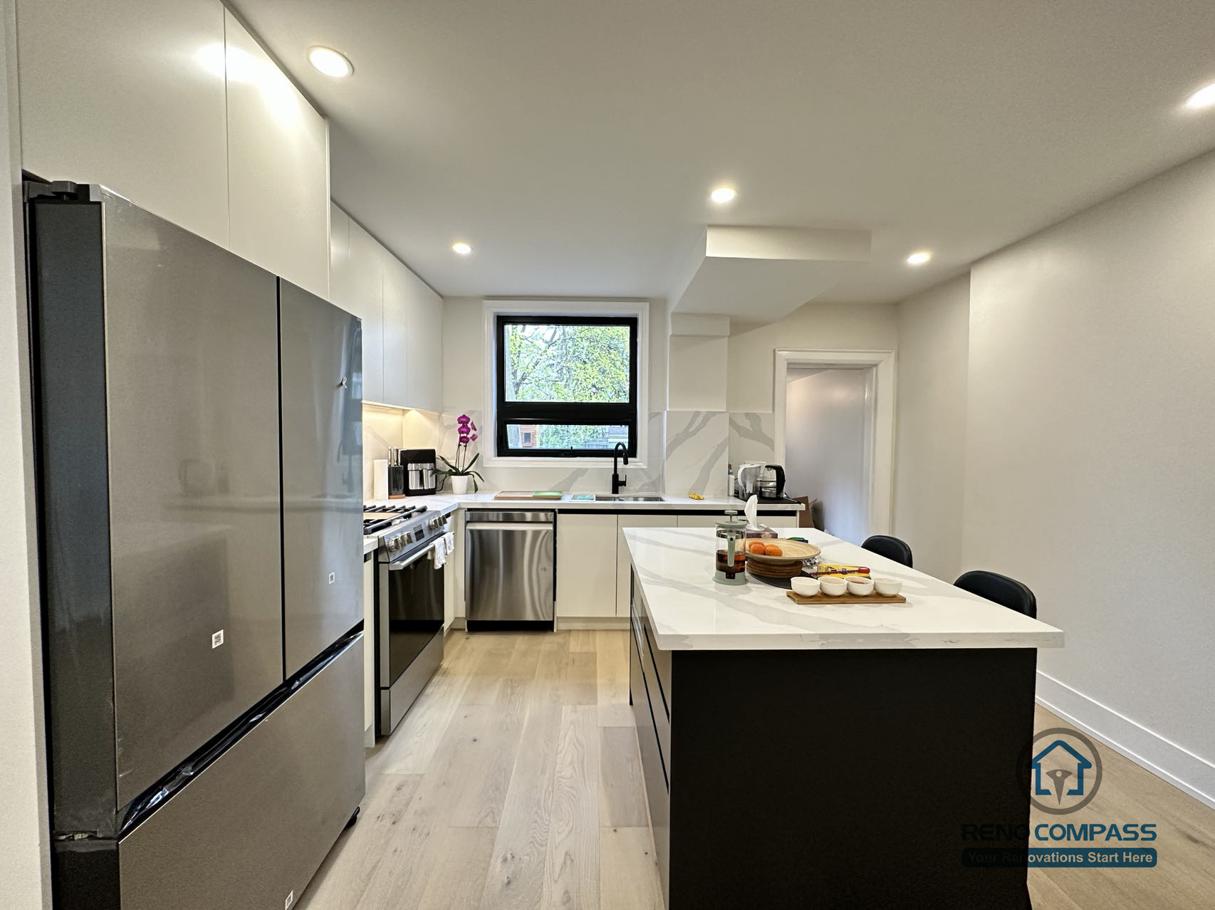 This Toronto interior renovation highlights a modern galley-style kitchen featuring a quartz island, stainless steel appliances, flat-panel cabinetry, recessed lighting, and wide-plank hardwood flooring. The efficient layout maximizes workspace and circulation, reflecting thoughtful planning and craftsmanship by a professional Toronto renovation company.