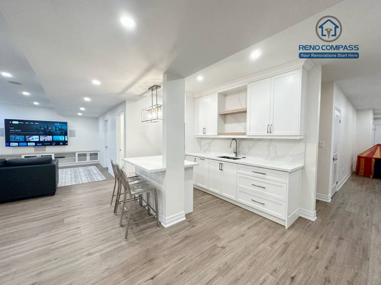 This Bradford basement renovation transforms the lower level into a bright, open-concept living and entertainment space. The layout integrates a custom wet bar with full-height cabinetry, quartz countertop, and built-in sink, creating a functional transition between the entertainment zone and seating area. Recessed LED lighting throughout the ceiling enhances brightness and eliminates the typical basement dark feel, while durable wood-look flooring provides warmth and continuity across the space. Structural columns were carefully incorporated into the design to maintain openness without compromising support. This basement renovation reflects Reno Compass’ approach to practical space planning, clean finishes, and long-term usability for modern family living.