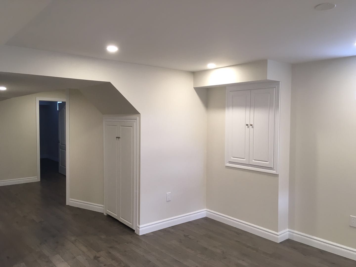 This Toronto basement renovation showcases a clean and functional living space with durable wood-look flooring, recessed pot lights, and custom built-in storage cabinets. The layout maximizes usable space under the staircase while maintaining a bright and open feel. Neutral wall tones and smart storage solutions create a versatile basement area suitable for everyday living or additional functional use.