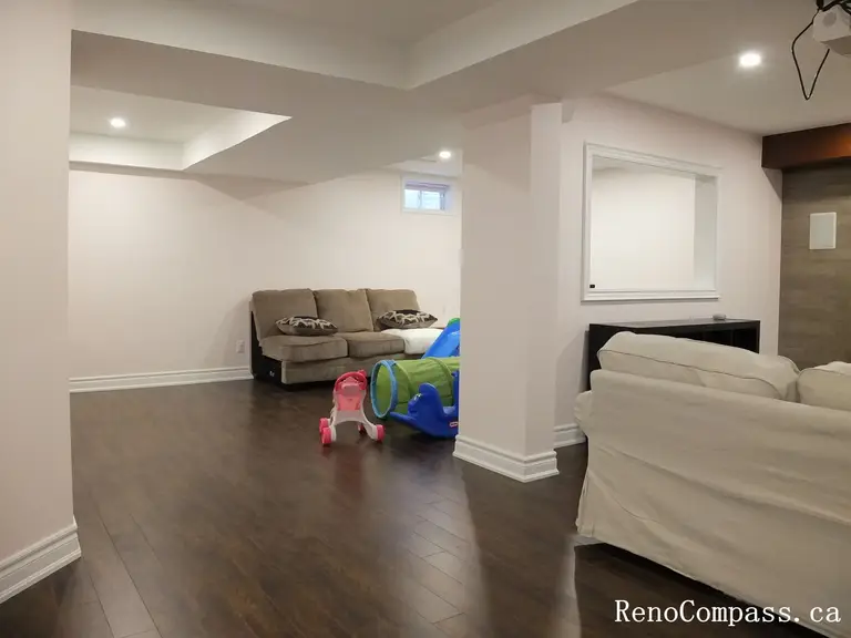 This Newmarket interior renovation showcases a bright and spacious finished basement designed for family living and relaxation. The space features dark hardwood flooring, recessed pot lights, structural columns, and a comfortable seating area that creates a warm and welcoming environment. The open layout allows the basement to function as a play area and lounge space, making it ideal for both adults and children. Completed by Reno Compass Inc., this renovation highlights practical space planning and modern basement finishing for everyday comfort.