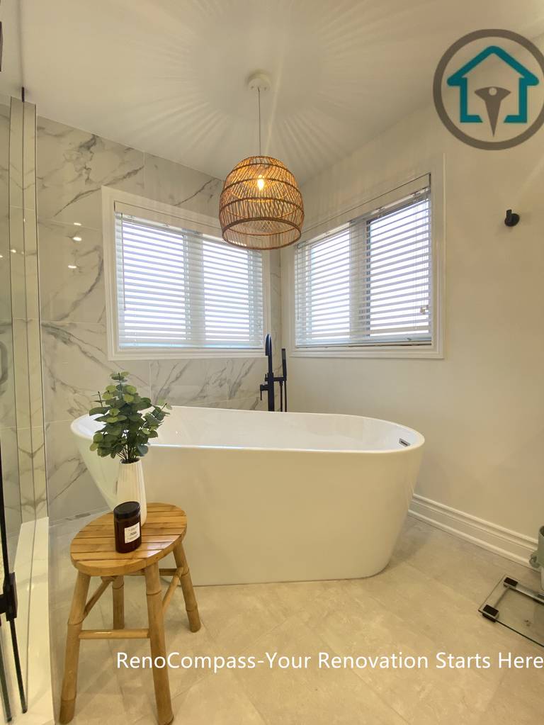 Elegant Oakville bathroom renovation featuring a freestanding bathtub positioned by corner windows, complemented by marble-look wall tiles, matte black fixtures, and a warm woven pendant light. This bathroom remodel blends natural light, soft textures, and spa-inspired comfort, completed by a trusted Toronto renovation company.