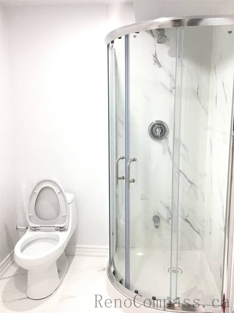 Modern basement bathroom renovation in Pickering featuring a curved glass shower enclosure with marble-style wall panels and a clean white toilet installation. The bright neutral finishes and compact layout maximize functionality while maintaining a sleek contemporary look. Professionally designed and completed by Reno Compass Inc., a trusted Toronto Renovation Company.