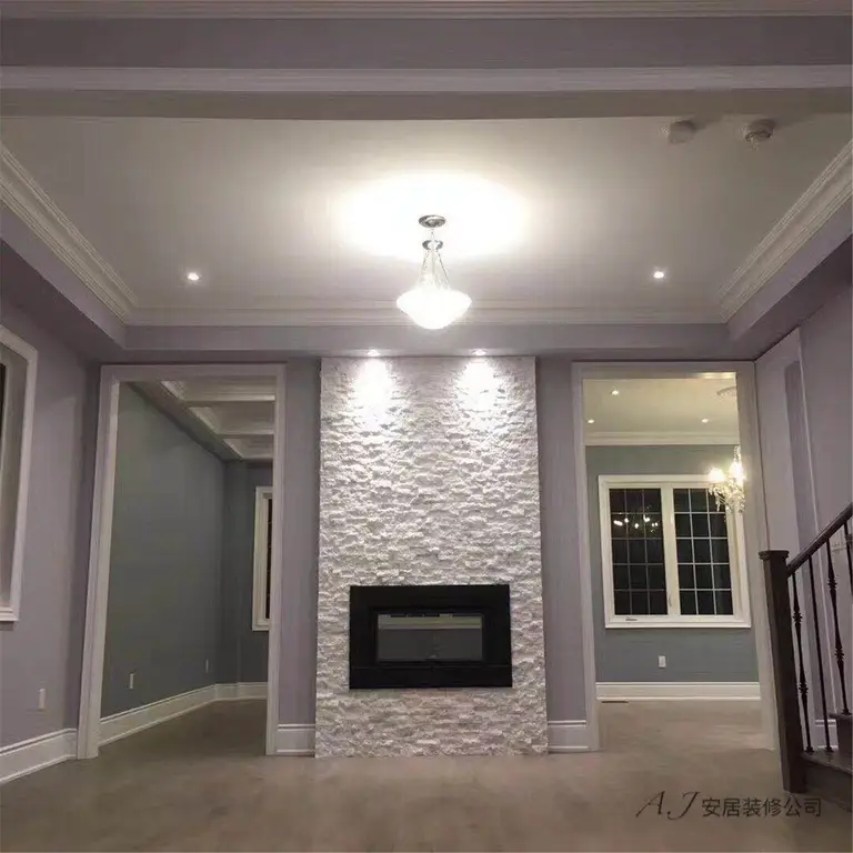 This Scarborough interior renovation showcases a modern living room centered around a textured stone fireplace feature wall. The space is enhanced with elegant crown molding, recessed ceiling lights, and neutral grey wall tones that create a calm and refined atmosphere. Large windows bring in natural light while the warm wood flooring adds balance and warmth to the overall design, highlighting a clean and contemporary interior renovation in Scarborough.