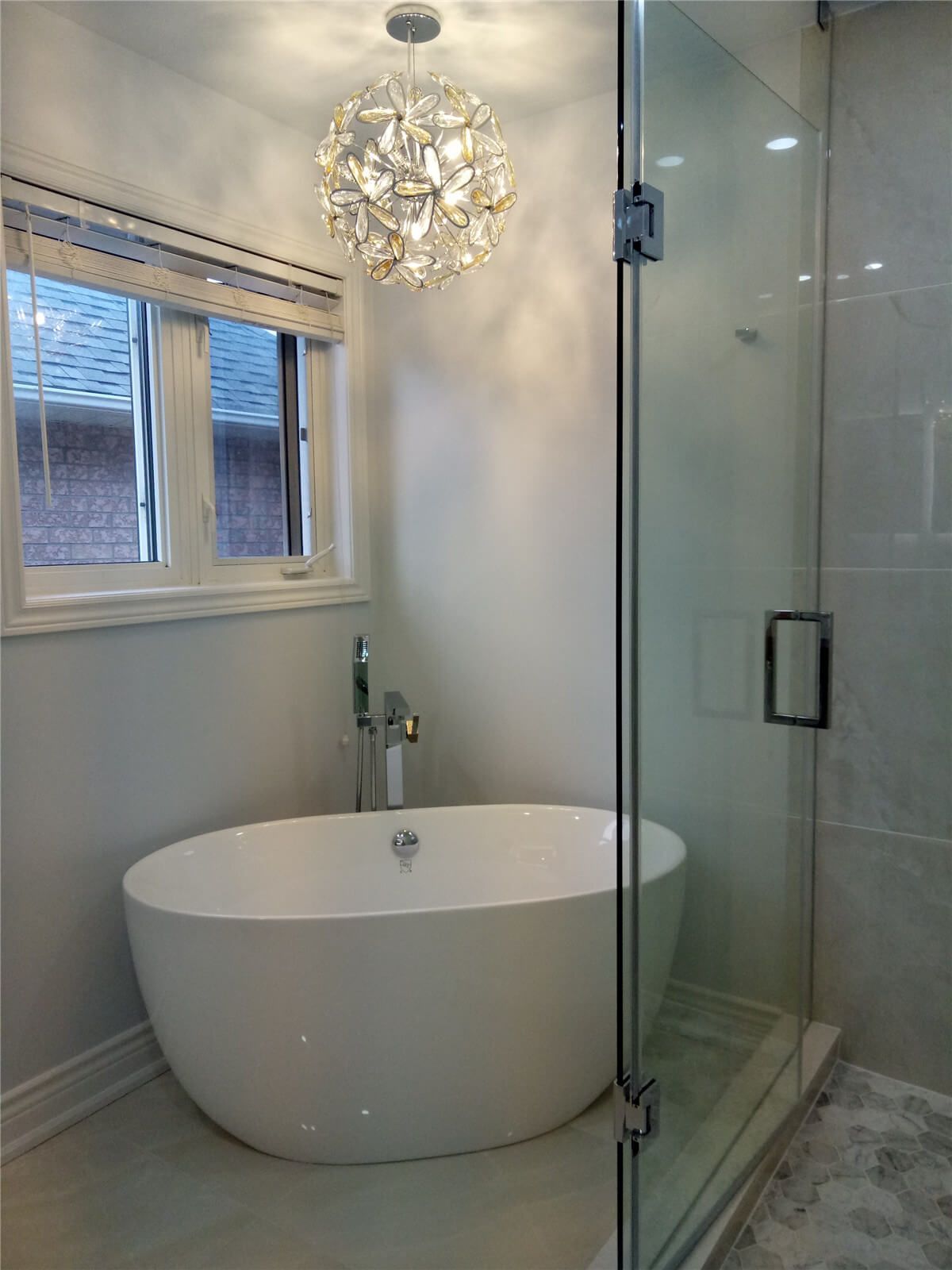 This Richmond Hill bathroom renovation features a modern freestanding bathtub paired with a frameless glass shower and elegant lighting fixture to create a relaxing, spa-inspired atmosphere. Large windows bring in natural light while neutral finishes and clean lines enhance the sense of space and comfort. The upgraded layout and high-quality fixtures deliver both functionality and visual appeal, transforming the bathroom into a luxurious retreat within this interior renovation project.