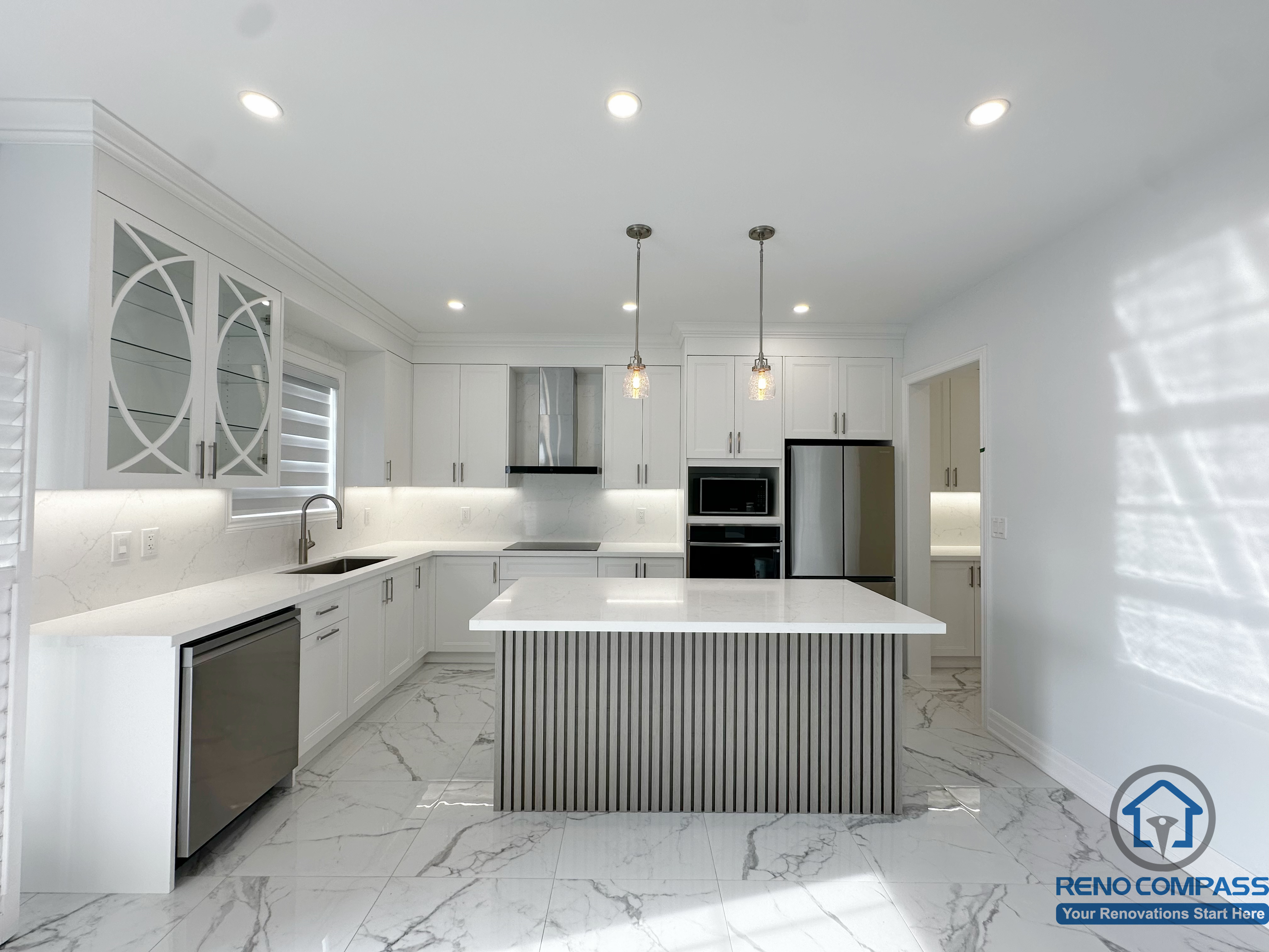 This image presents the main view of a modern kitchen renovation in a Markham house. The space features custom white cabinetry, a large central island with vertical panel detailing, marble-style flooring, integrated stainless steel appliances, and balanced lighting that enhances brightness, functionality, and everyday usability.