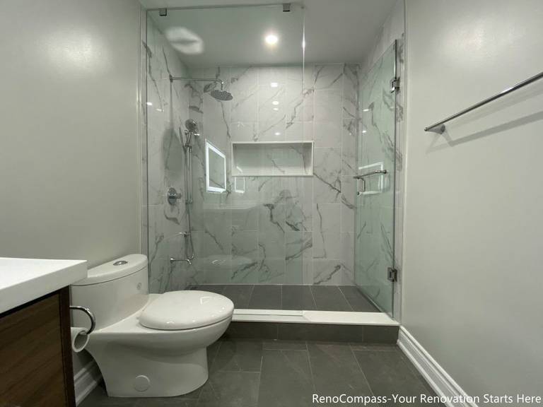 This Oakville bathroom renovation showcases a modern walk-in glass shower with marble-look wall tiles, built-in shower niche, and sleek frameless enclosure for a clean contemporary design. The space also features a custom wood vanity with quartz countertop, LED backlit mirror, and upgraded fixtures that enhance both functionality and visual appeal. Completed by a professional Toronto renovation company, this bathroom upgrade focuses on maximizing brightness, storage, and comfort while delivering a stylish and durable modern bathroom space.
