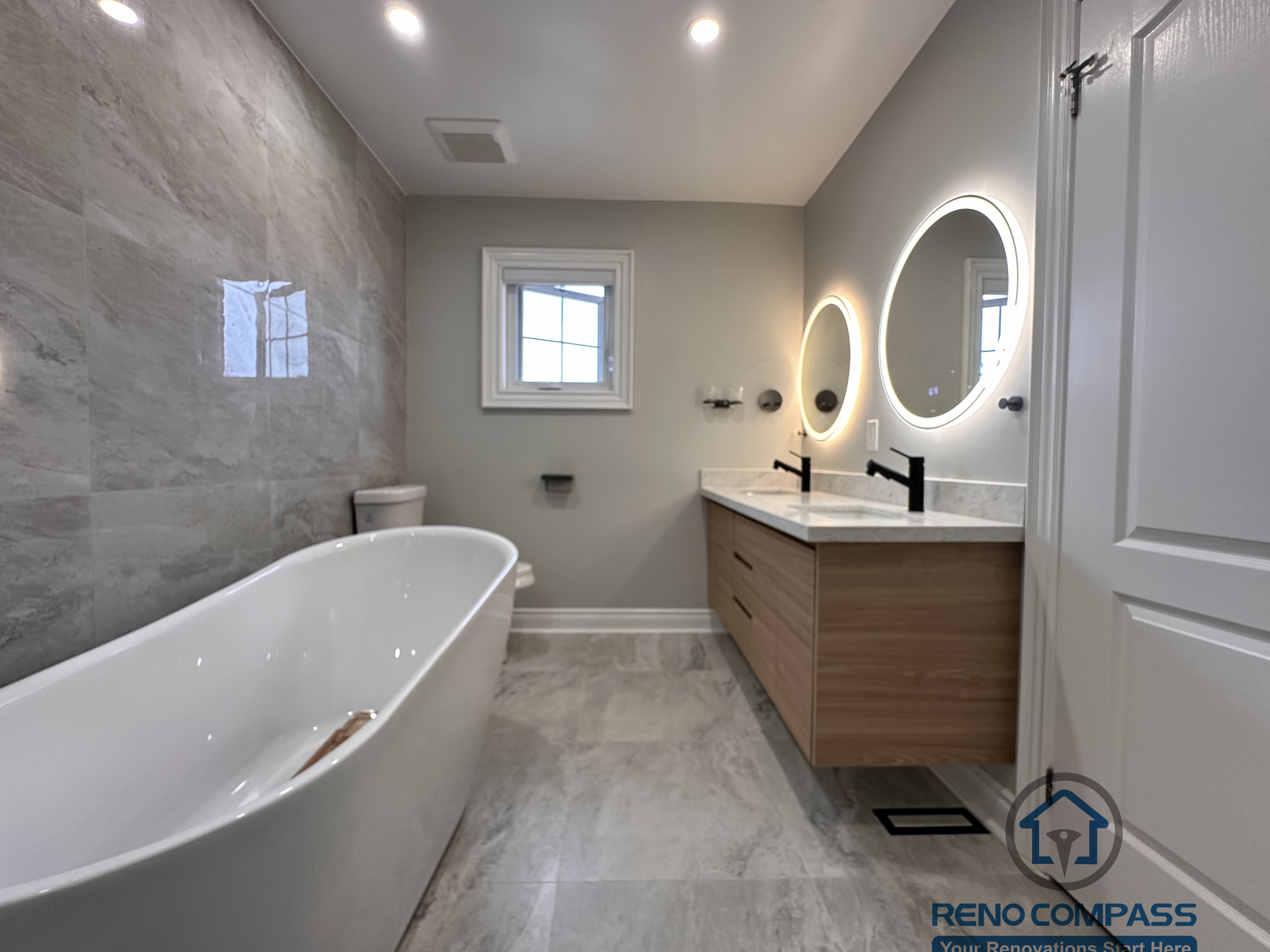 This Markham bathroom renovation features a custom wood-tone vanity with a quartz countertop, round LED backlit mirror, and matte black fixtures. The layout combines a glass-enclosed shower and bathtub with a practical vanity and toilet arrangement, creating a clean and well-balanced space. Designed and built by a professional Toronto renovation company, Reno Compass Inc., this bathroom emphasizes functionality, durability, and timeless design.