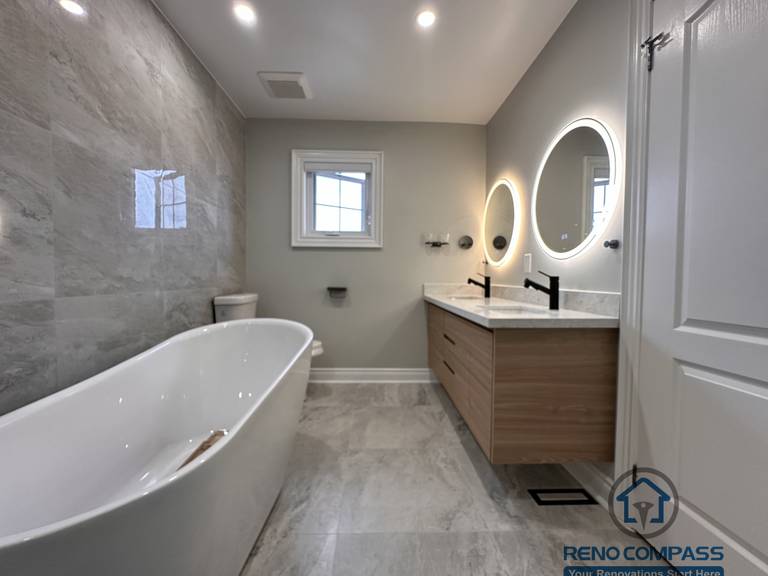 This Markham bathroom renovation features a custom wood-tone vanity with a quartz countertop, round LED backlit mirror, and matte black fixtures. The layout combines a glass-enclosed shower and bathtub with a practical vanity and toilet arrangement, creating a clean and well-balanced space. Designed and built by a professional Toronto renovation company, Reno Compass Inc., this bathroom emphasizes functionality, durability, and timeless design.
