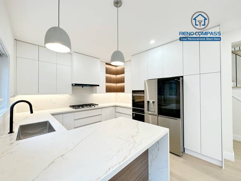 This image presents a modern kitchen renovation in Richmond Hill featuring full-height custom white cabinetry, a quartz waterfall island with wood panel detailing, and integrated stainless steel appliances. The design includes a built-in gas cooktop, minimalist pendant lights, recessed ceiling lighting, and light hardwood flooring for a clean, contemporary look. Large windows and patio access enhance natural light and openness. Completed by a professional Toronto Renovation Company, this kitchen renovation reflects refined craftsmanship, efficient layout planning, and modern family-focused design.