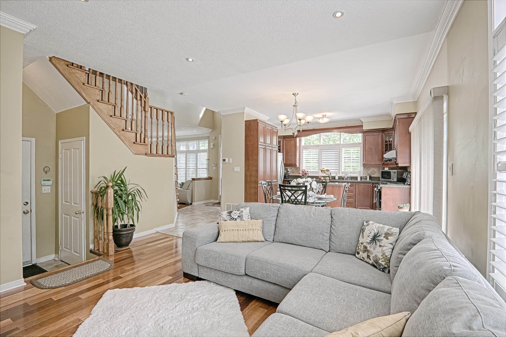 This open-concept living room and kitchen layout features warm hardwood flooring, soft neutral tones, and a seamless connection between living, dining, and kitchen spaces. The cozy sectional sofa, natural lighting, and classic wood staircase enhance both comfort and visual flow throughout the home. Designed for modern family living, this space reflects a perfect balance of functionality and warmth. A great example of open-concept home renovation and interior design inspiration for Toronto and GTA homeowners.