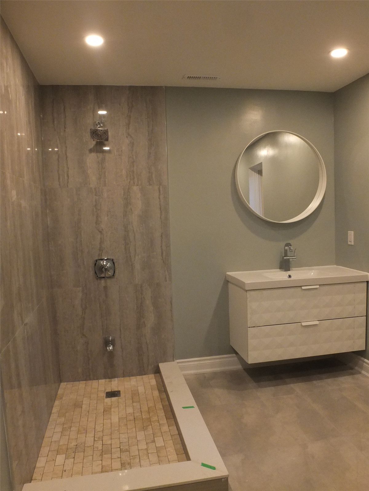 This image features a modern bathroom renovation completed as part of a Mississauga interior renovation project. The design includes a walk-in tiled shower with full-height wall tiles, recessed ceiling lighting, and a sleek floating vanity paired with a round mirror. Neutral wall colors and clean lines create a bright, minimalist atmosphere while maximizing functionality and comfort. Completed by an experienced Toronto renovation company, this bathroom renovation improves daily usability, moisture resistance, and overall interior value for homeowners in Mississauga and the Greater Toronto Area.