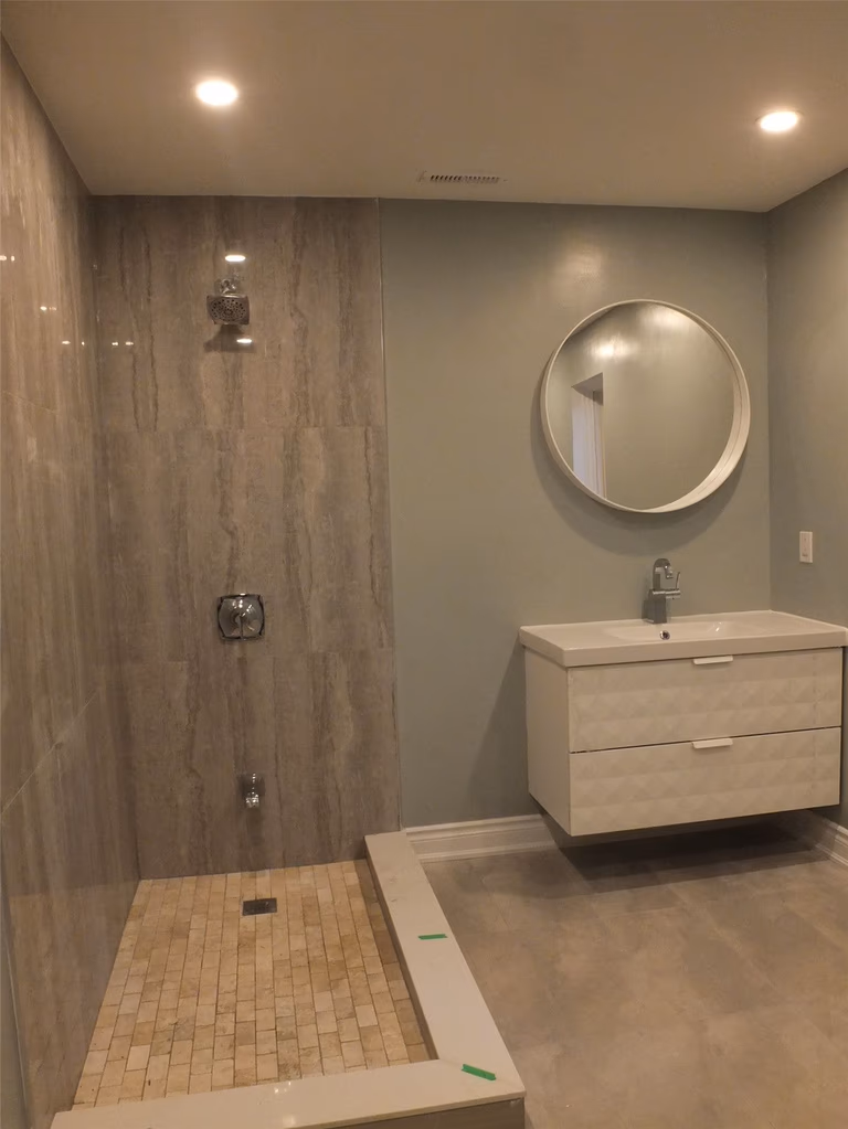 This image features a modern bathroom renovation completed as part of a Mississauga interior renovation project. The design includes a walk-in tiled shower with full-height wall tiles, recessed ceiling lighting, and a sleek floating vanity paired with a round mirror. Neutral wall colors and clean lines create a bright, minimalist atmosphere while maximizing functionality and comfort. Completed by an experienced Toronto renovation company, this bathroom renovation improves daily usability, moisture resistance, and overall interior value for homeowners in Mississauga and the Greater Toronto Area.