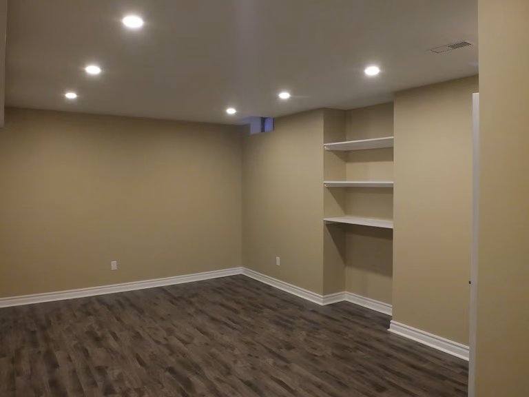 This Vaughan basement renovation showcases a clean and functional finished space designed by a professional Toronto Renovation Company. The layout features modern recessed pot lighting, durable wood-look flooring, and custom built-in shelving that provides both storage and decorative display options. Neutral wall tones and thoughtful lighting placement create a warm and inviting environment, ideal for a family lounge, entertainment room, or home office. The finished basement enhances everyday living while adding long-term property value for homeowners across the GTA.