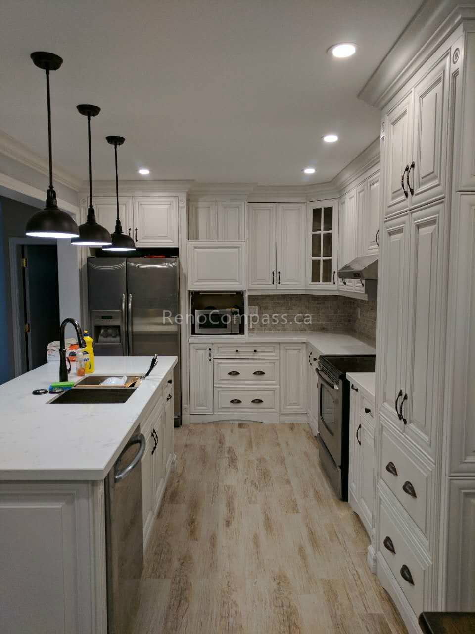 This Vaughan kitchen renovation by Reno Compass Inc. showcases custom white shaker cabinets, a large quartz island with seating, and sleek black pendant lighting. The open-concept layout enhances storage, workflow, and natural light, while stainless steel appliances and warm wood flooring create a clean, modern, and highly functional kitchen designed for daily use and entertaining.