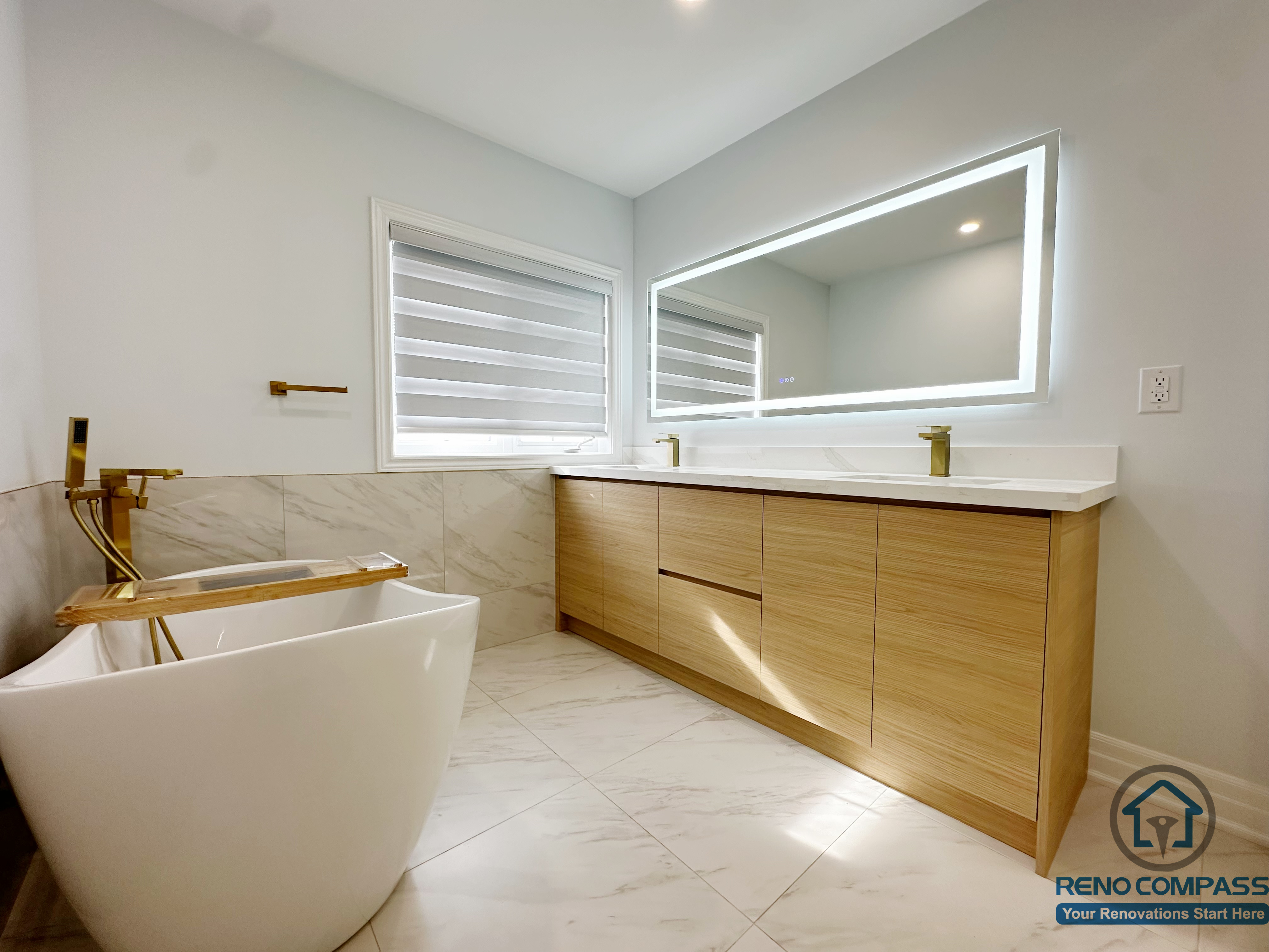 This image features a modern bathroom renovation in a Markham house, showcasing a freestanding soaking tub with brushed gold fixtures, a custom wood vanity with integrated storage, and a large LED backlit mirror. Marble-style floor and wall tiles, combined with natural wood tones, create a clean, spa-inspired bathroom design with a warm and contemporary feel.