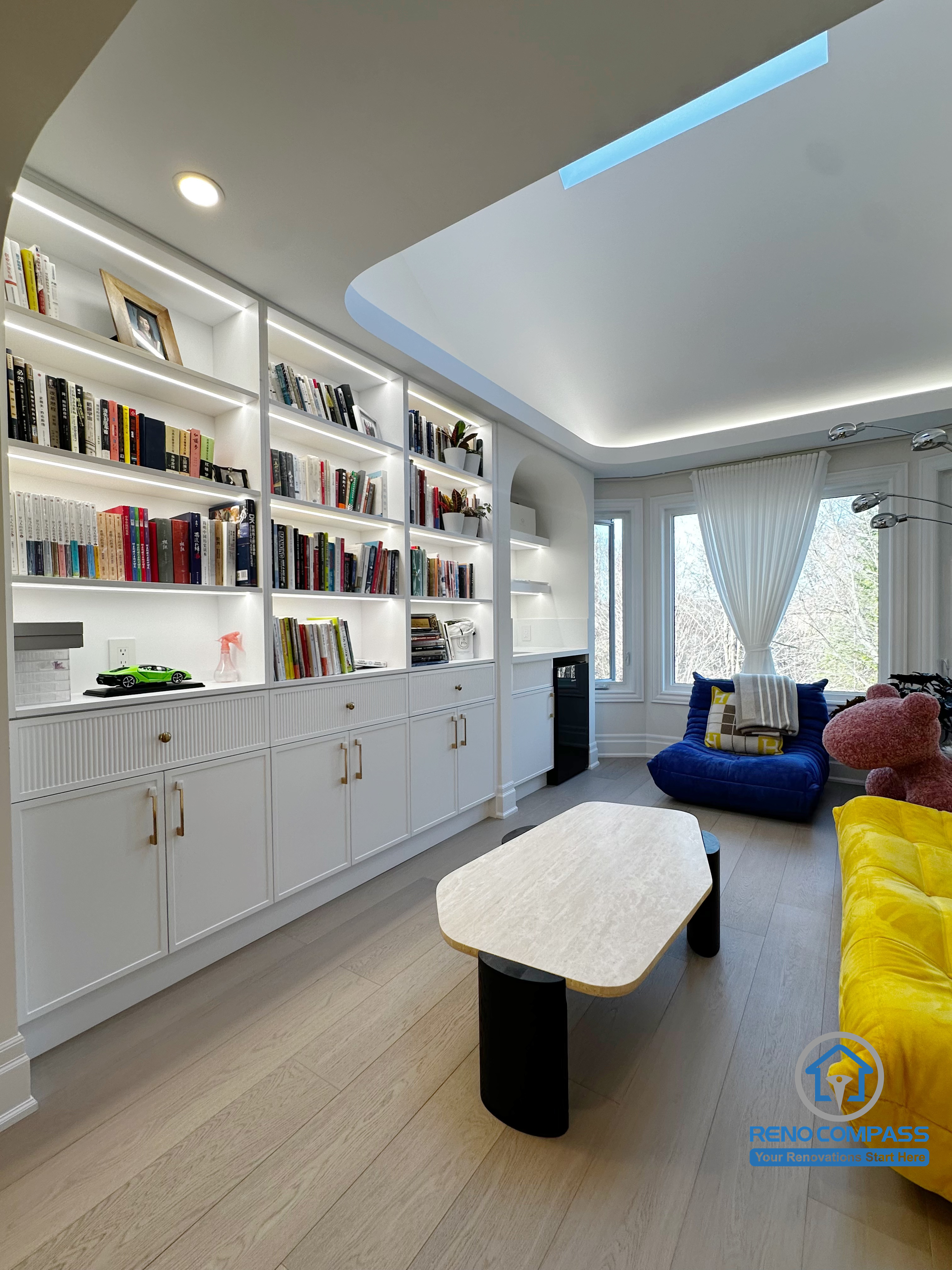 This area of the Markham interior renovation was designed as a calm, functional lounge that combines custom storage with everyday comfort. Full-height built-in shelving along one wall provides organized space for books and personal items while maintaining a clean, integrated appearance within the room.

The ceiling design features recessed lighting and soft indirect illumination, creating an even and inviting atmosphere without visual clutter. Light wood flooring helps balance the white cabinetry and bright furnishings, ensuring the space feels warm rather than overly minimal.

Designed for reading, relaxation, and quiet family time, this lounge area reflects a thoughtful approach to interior planning—where storage, lighting, and comfort work together to support daily living.