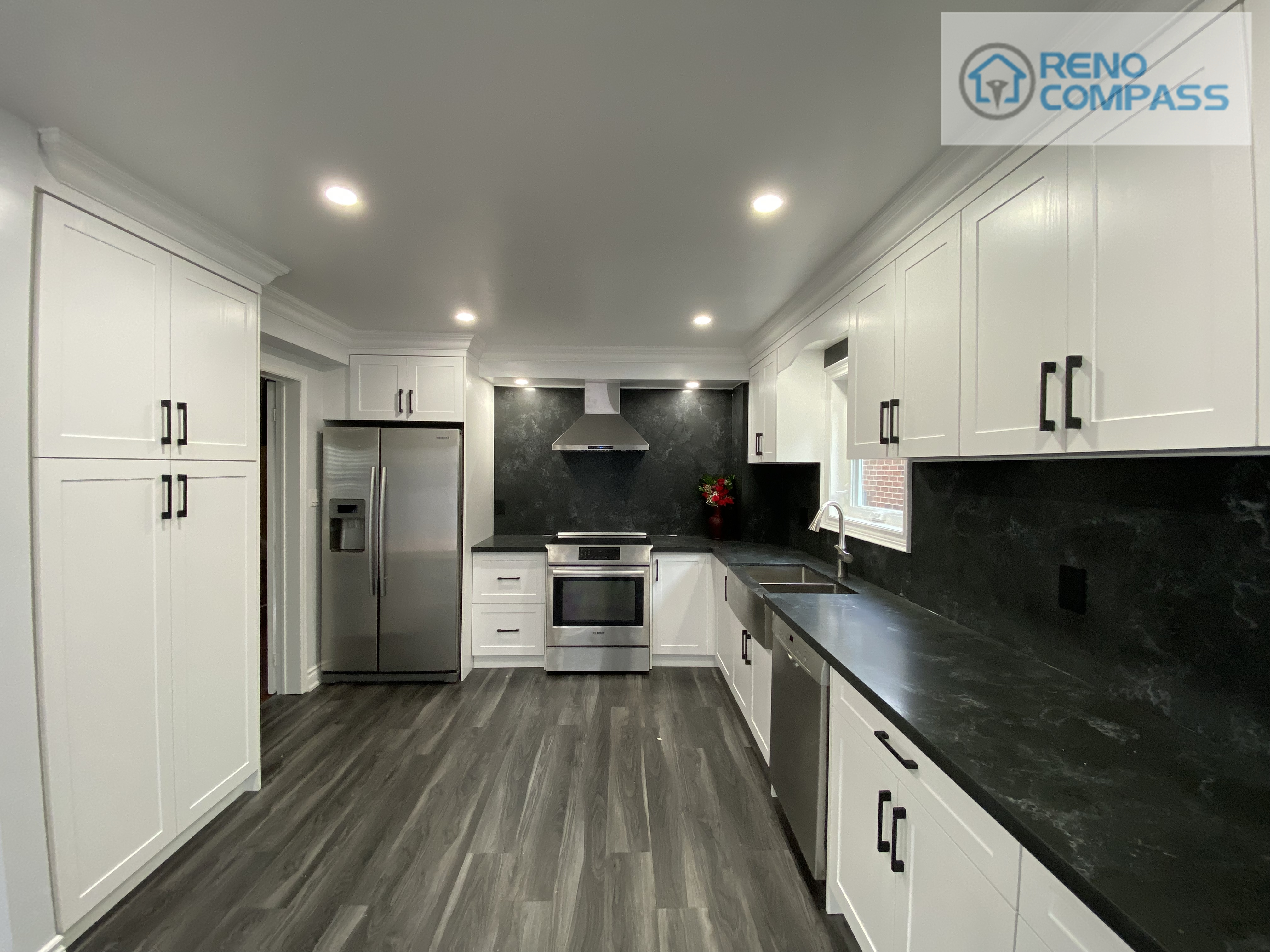 Wide-angle view of a modern kitchen interior renovation in Pickering, featuring full-height white shaker cabinetry, black stone countertops and backsplash, stainless steel appliances, recessed ceiling lighting, and durable vinyl plank flooring. This clean and functional kitchen layout was designed and built by Reno Compass Inc., delivering a bright, streamlined space optimized for everyday use.