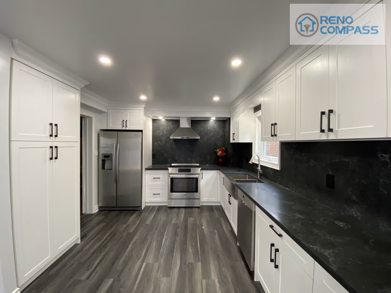 Wide-angle view of a modern kitchen interior renovation in Pickering, featuring full-height white shaker cabinetry, black stone countertops and backsplash, stainless steel appliances, recessed ceiling lighting, and durable vinyl plank flooring. This clean and functional kitchen layout was designed and built by Reno Compass Inc., delivering a bright, streamlined space optimized for everyday use.