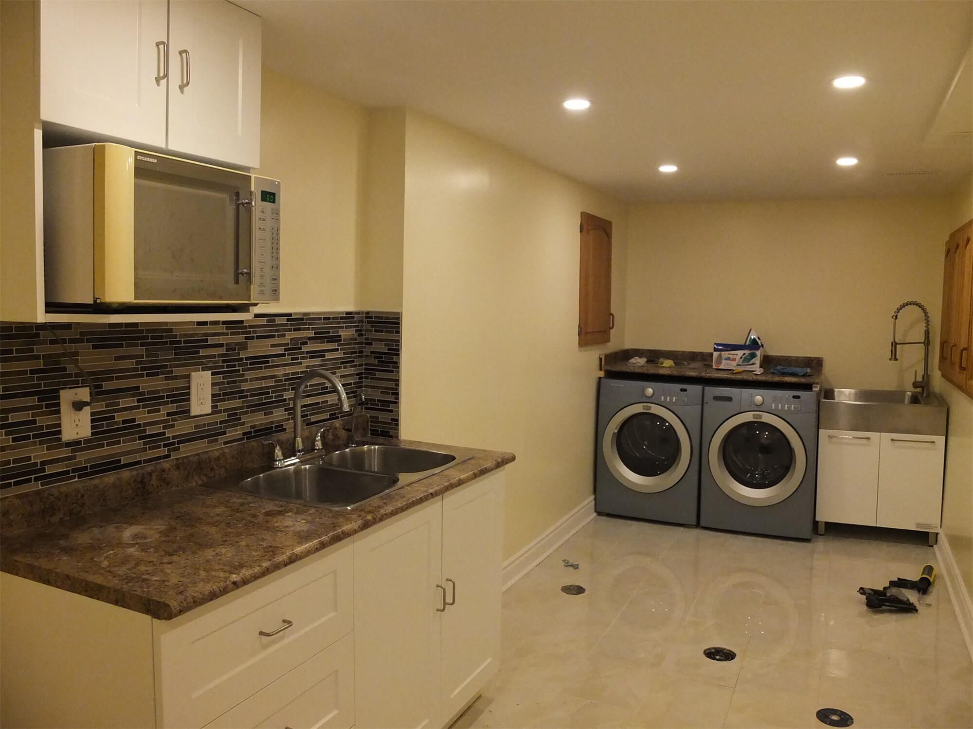 This Oshawa basement renovation features a newly installed kitchenette and laundry area designed for convenience and functionality. The space includes modern white cabinetry, a durable laminate countertop, mosaic tile backsplash, and recessed pot lighting for a clean and practical layout. A stainless-steel sink and built-in microwave enhance usability, while the dedicated washer and dryer setup provides added efficiency for everyday living. The polished tile flooring ensures durability and easy maintenance. Completed by a professional Toronto Renovation Company, this project demonstrates thoughtful planning and quality craftsmanship in transforming a basement into a fully functional living space.