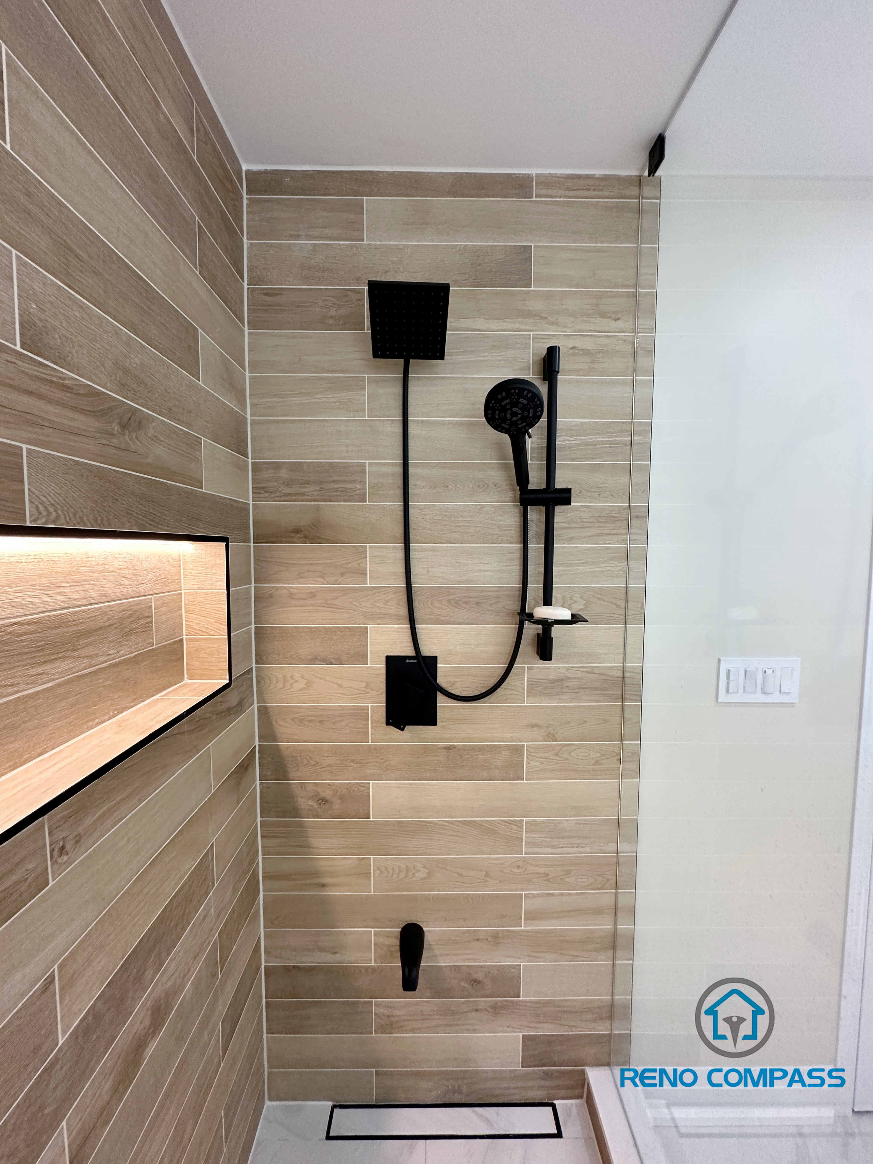 Contemporary bathroom renovation in Vaughan showcasing a sleek matte black shower system with a square rain shower head and handheld fixture. The shower walls are finished with wood-look porcelain tiles installed in a horizontal layout, creating a warm and modern spa-inspired aesthetic. Paired with a frameless glass shower enclosure, this design highlights clean lines, premium materials, and precise installation by an experienced Toronto renovation company.