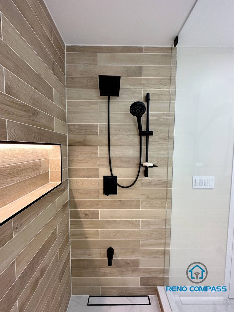 Contemporary bathroom renovation in Vaughan showcasing a sleek matte black shower system with a square rain shower head and handheld fixture. The shower walls are finished with wood-look porcelain tiles installed in a horizontal layout, creating a warm and modern spa-inspired aesthetic. Paired with a frameless glass shower enclosure, this design highlights clean lines, premium materials, and precise installation by an experienced Toronto renovation company.