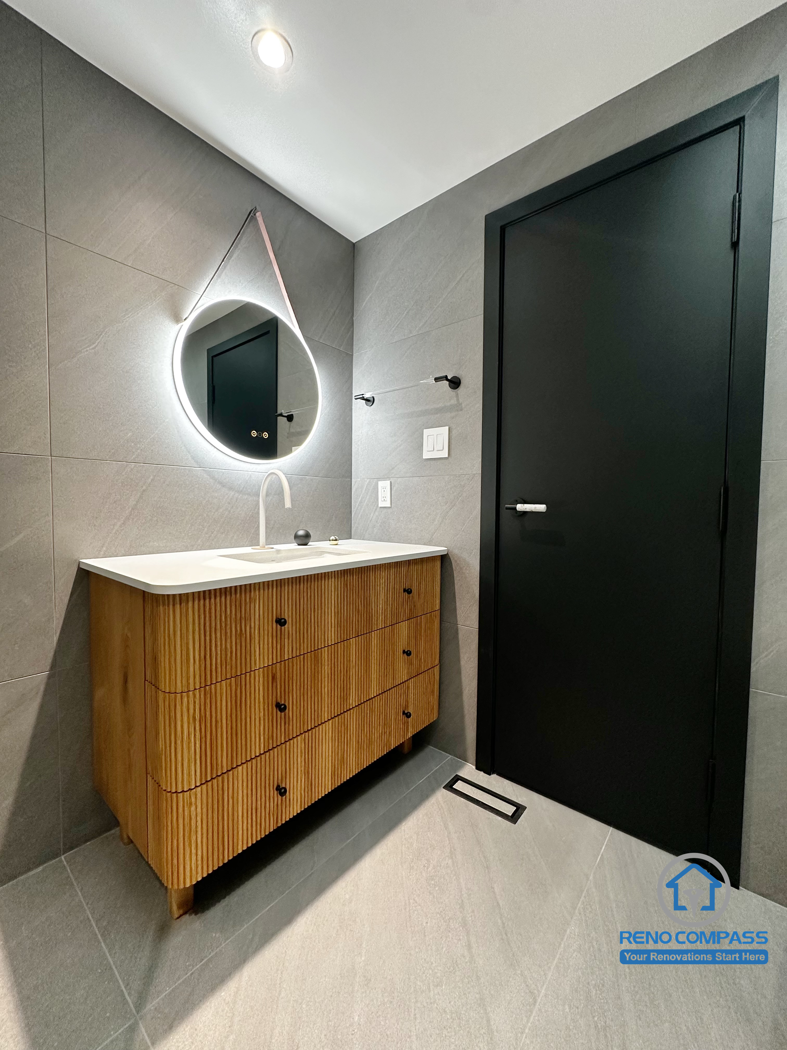 This view showcases the bathroom vanity area in a Richmond Hill renovation, featuring a custom wood-textured vanity, an integrated sink, and a sculptural backlit mirror. The matte black door and minimalist hardware create contrast against the large-format grey tiles, enhancing depth and spatial clarity. Designed and completed by Reno Compass, this bathroom reflects a clean, modern renovation approach in the Greater Toronto Area.