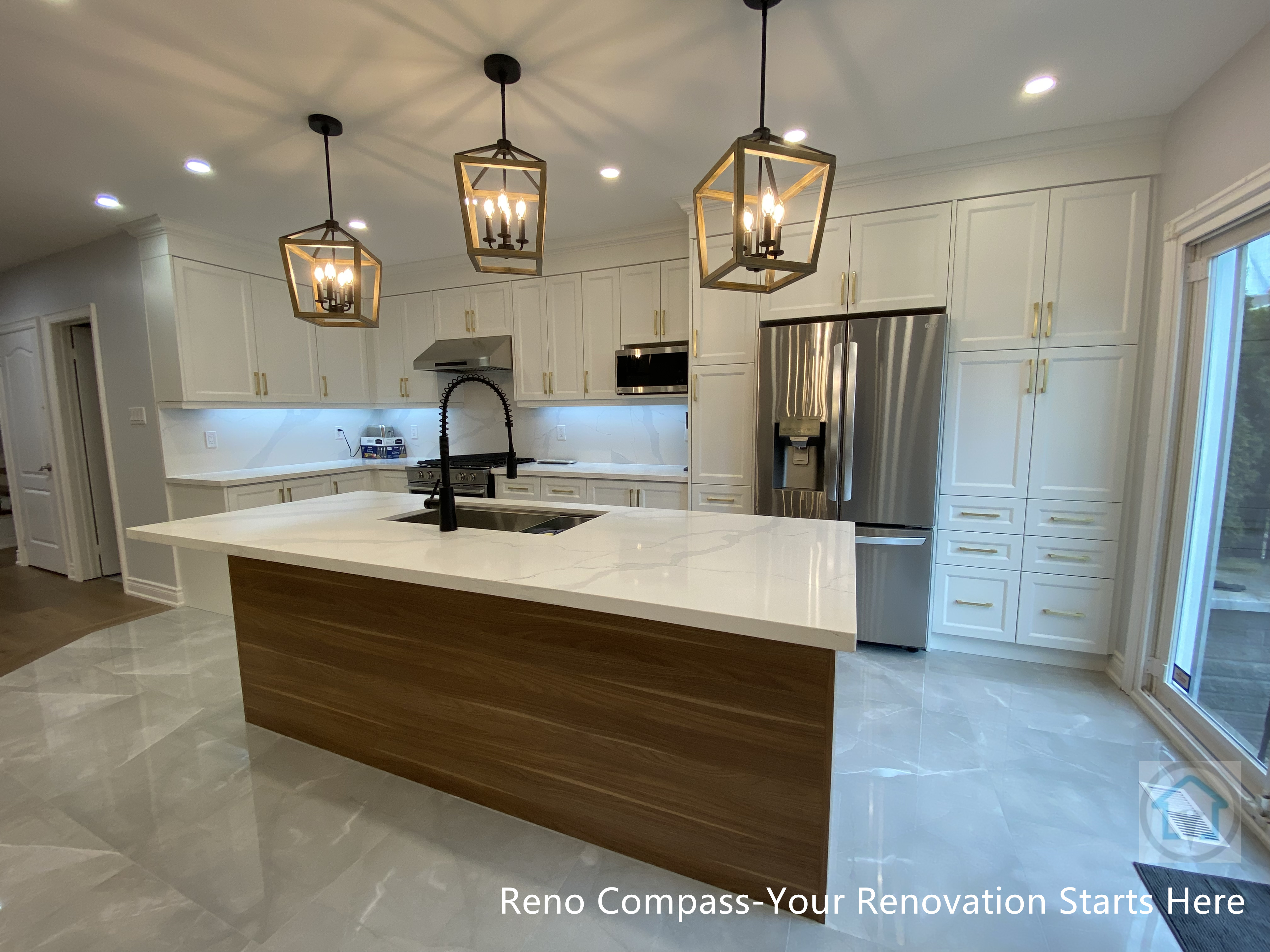 This image highlights a Newmarket interior renovation featuring a long quartz kitchen island with warm wood base cabinetry, designed to anchor the open-concept layout. The linear arrangement of lantern-style pendant lights creates strong visual rhythm and enhances the sense of depth within the space. White shaker cabinets, integrated stainless steel appliances, and glossy marble-look flooring work together to reflect light and elevate the overall brightness. A high-arc faucet and built-in sink add both functionality and modern appeal. Completed by a Toronto renovation company, this kitchen renovation emphasizes clean lines, balanced proportions, and seamless flow between cooking, dining, and outdoor access.