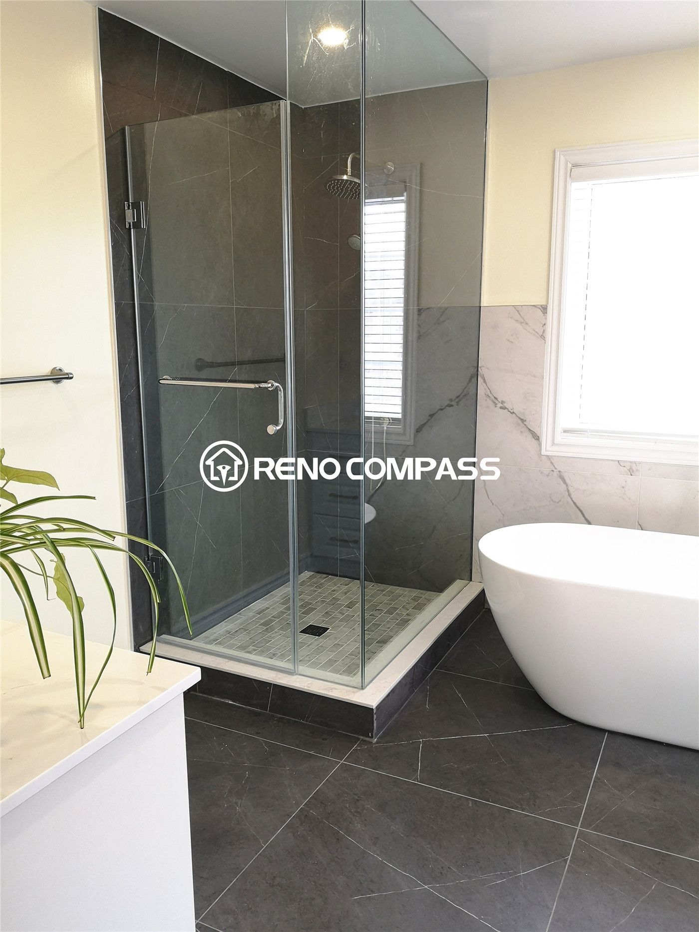 This modern Toronto bathroom renovation features a frameless glass shower paired with a stylish freestanding bathtub. Dark porcelain tiles and marble wall accents create a balanced contrast for a sleek, contemporary feel. Designed by Reno Compass, this remodel enhances both luxury and livability with elegant finishes and thoughtful layout.