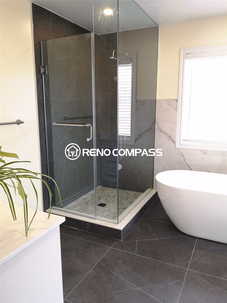 This modern Toronto bathroom renovation features a frameless glass shower paired with a stylish freestanding bathtub. Dark porcelain tiles and marble wall accents create a balanced contrast for a sleek, contemporary feel. Designed by Reno Compass, this remodel enhances both luxury and livability with elegant finishes and thoughtful layout.