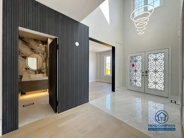 This image showcases a dramatic entryway from a Markham interior renovation, where architectural detailing and material contrast define the first impression of the home. A full-height vertical slat feature wall in a dark finish anchors the space, providing visual depth while framing the entrance to the powder room.

The powder room is subtly revealed behind the feature wall, finished with stone-pattern wall surfaces, a floating vanity, and a round backlit mirror that adds warmth and softness to the darker tones. Integrated lighting beneath the vanity enhances the sense of refinement and spatial layering.

The double-height foyer allows natural light to flow through decorative front doors, complemented by a sculptural modern chandelier that emphasizes vertical openness. The transition between polished tile flooring at the entry and light wood flooring beyond creates a clear yet seamless zoning between public and private areas.

This design reflects Reno Compass’s approach to modern interior renovation—where entryways are treated as architectural statements that balance functionality, privacy, and visual impact in Markham homes.