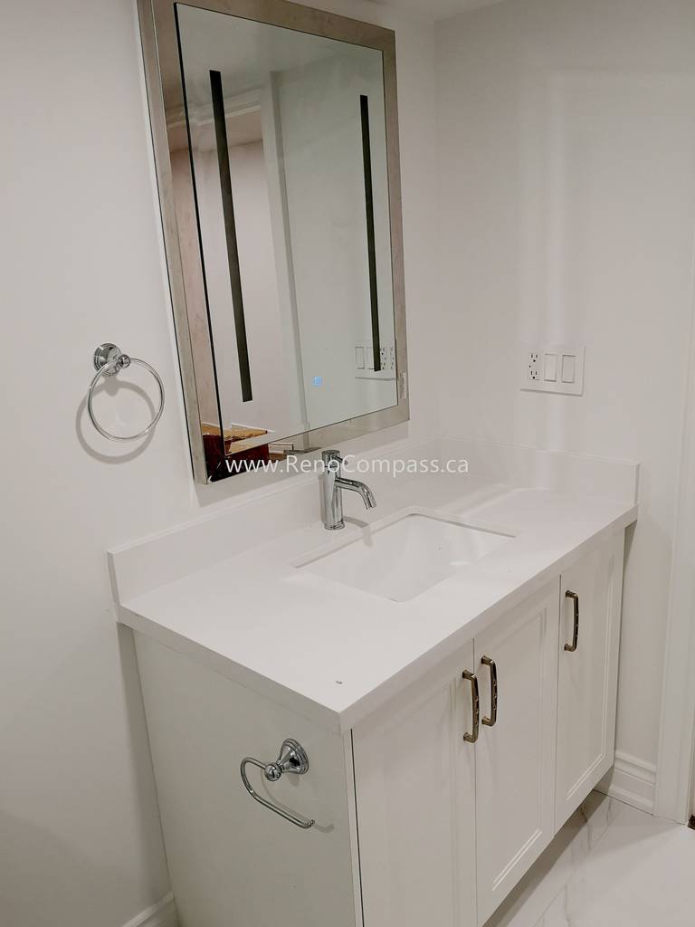 This Vaughan basement renovation features a modern bathroom vanity installation with a sleek white quartz countertop, under-mount sink, and contemporary chrome faucet. The clean white cabinetry provides both storage and a minimalist aesthetic, perfectly matching the bright and modern basement design. Completed by Reno Compass Inc., this basement bathroom upgrade enhances functionality while maintaining a stylish and cohesive interior renovation finish.